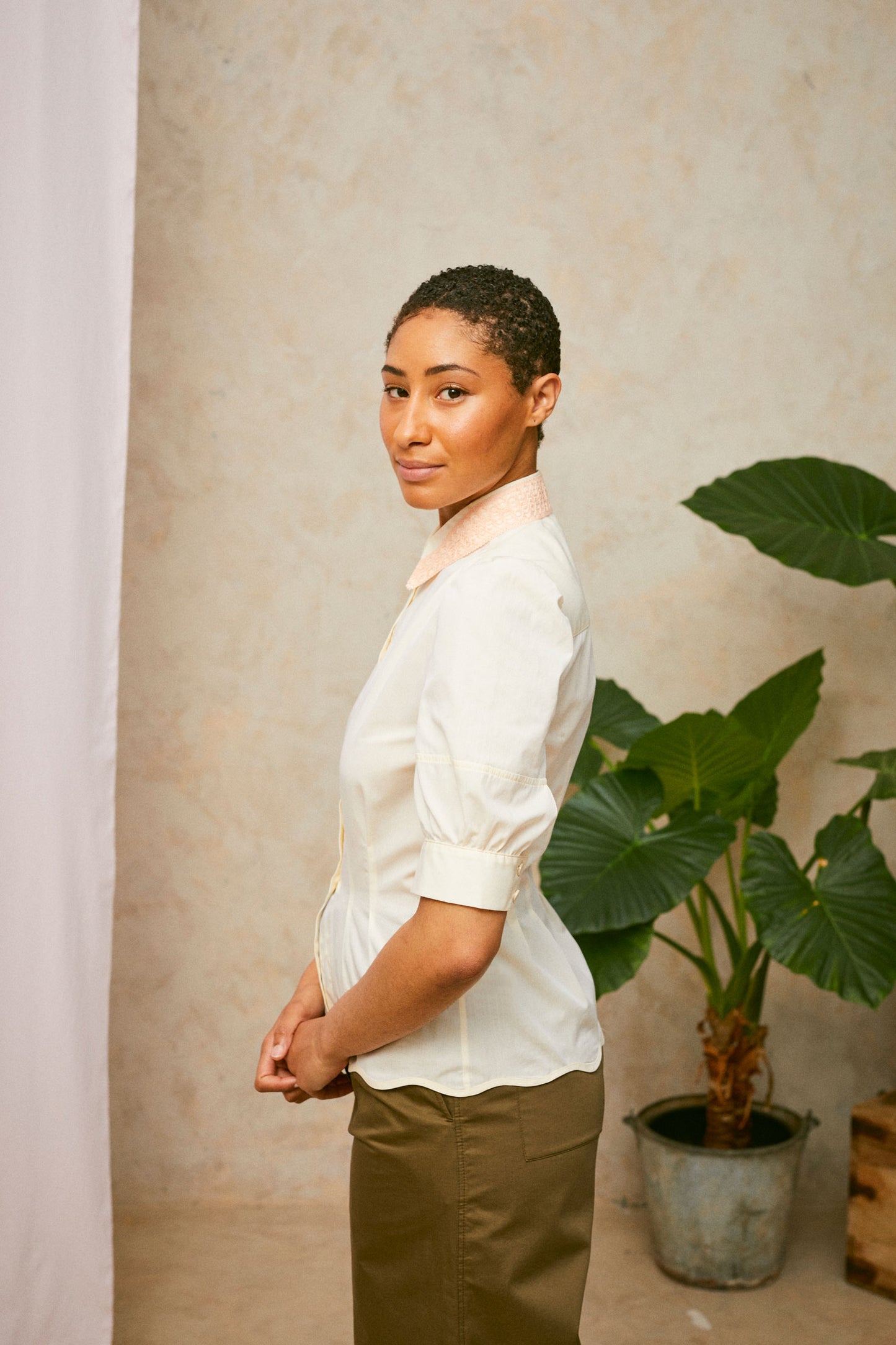Profile view of model wearing Saywood's pastel yellow Joni puff sleeve blouse with orange lace collar and scalloped hem. Worn with khaki Amelia wide leg trousers. A plant and drop of pink fabric can be seen in the background. 