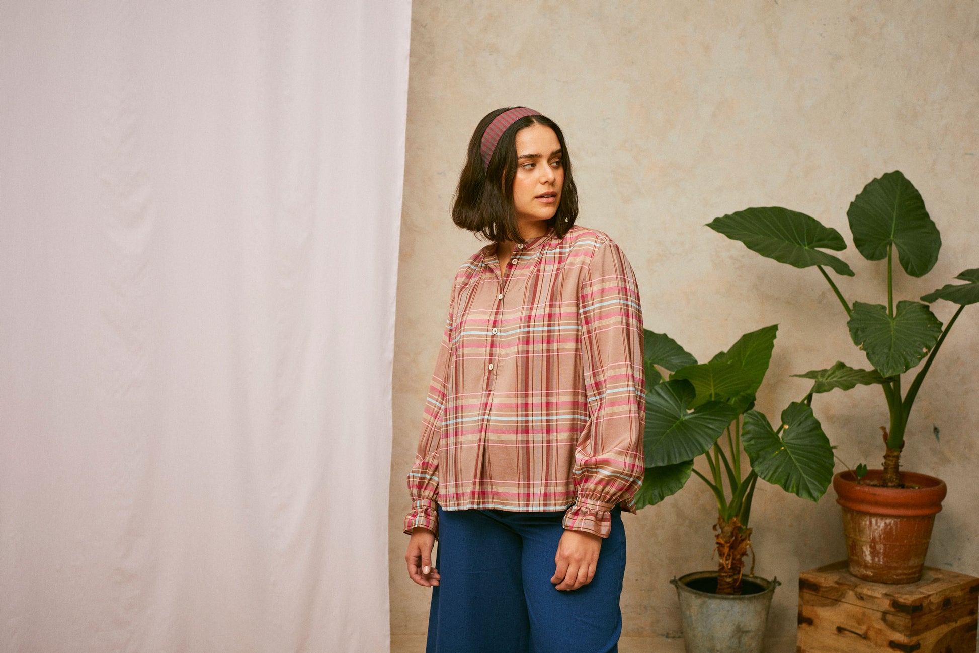 Model stands looking to the side. She wears Saywood's pink check shirt, the Marie A-Line Blouse, with natural indigo Amelia wide leg trousers. The red check Heidi Headband is worn in her her. A plant and drop of pink fabric can be seen in the background.