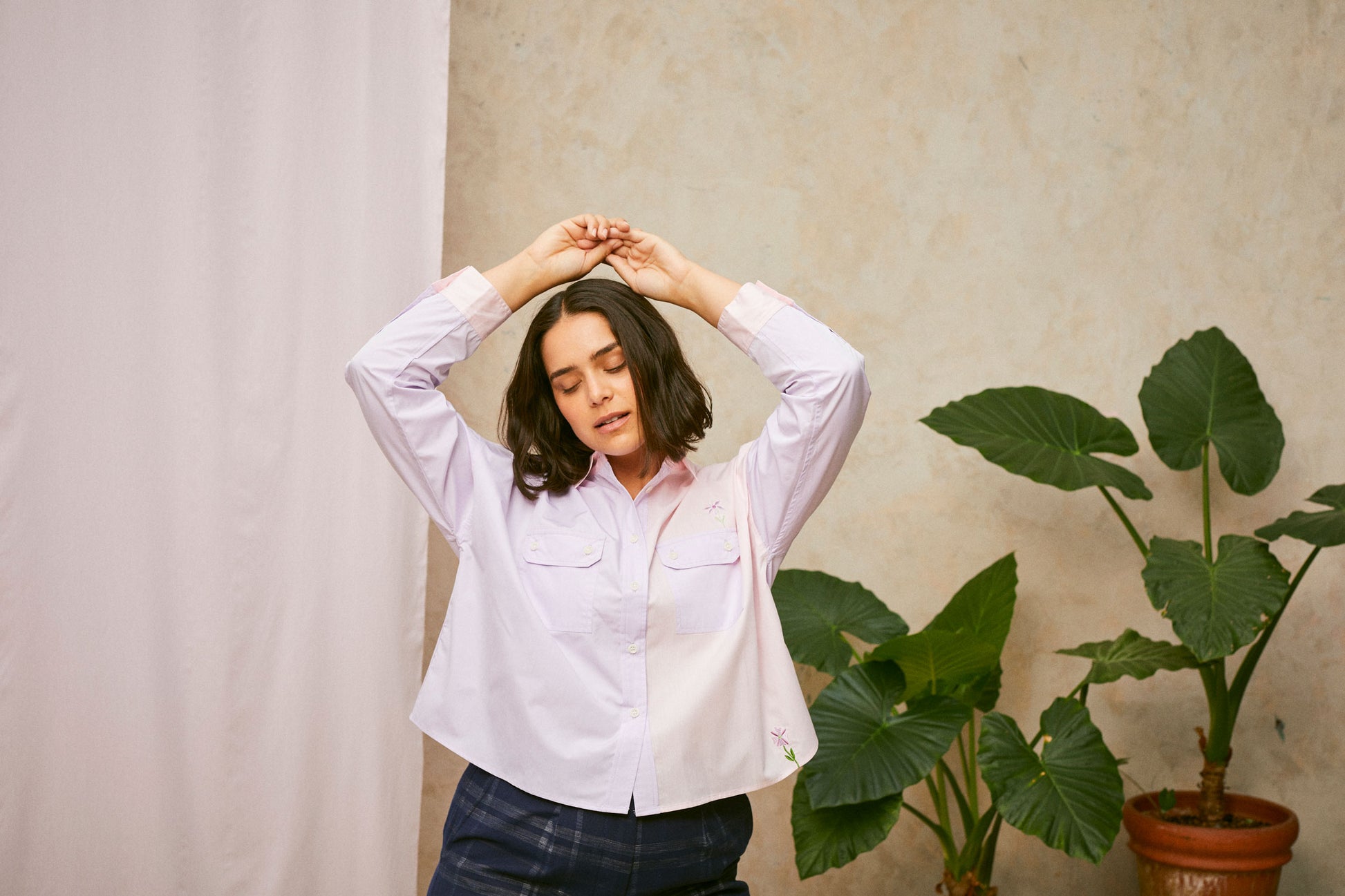 Model wears the Jules Utility Shirt: colourblocked pink and lilac shirt with a boxy silhouette. Jules Shirt from Saywood with embroidered flower detail and utility style pockets. Worn with a navy check trouser, she stands in front of a pink fabric drop and a plant, with her arms above her head.