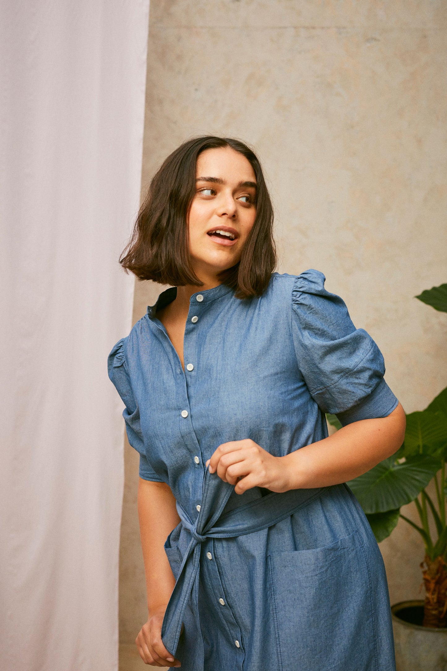 Close up of model wearing Saywood's Rosa denim shirtdress in light wash Japanese denim; a long line style with patch pockets and ruffles. She pulls at the belt tied round the waist whilst looking to the side. A plant and drop of pink fabric can be seen in the background.