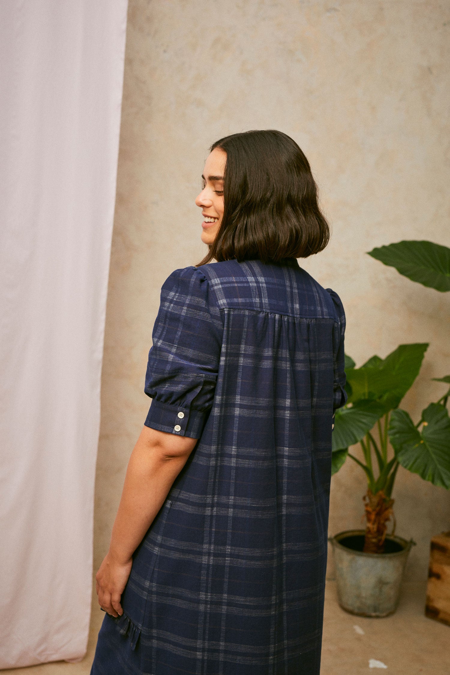 Cropped back view shot of model wearing Saywood's Rosa puff sleeve shirtdress in navy check cotton; gathers can be seen on the back of the dress from the yoke seam. A plant and drop of pink fabric can be seen in the background.