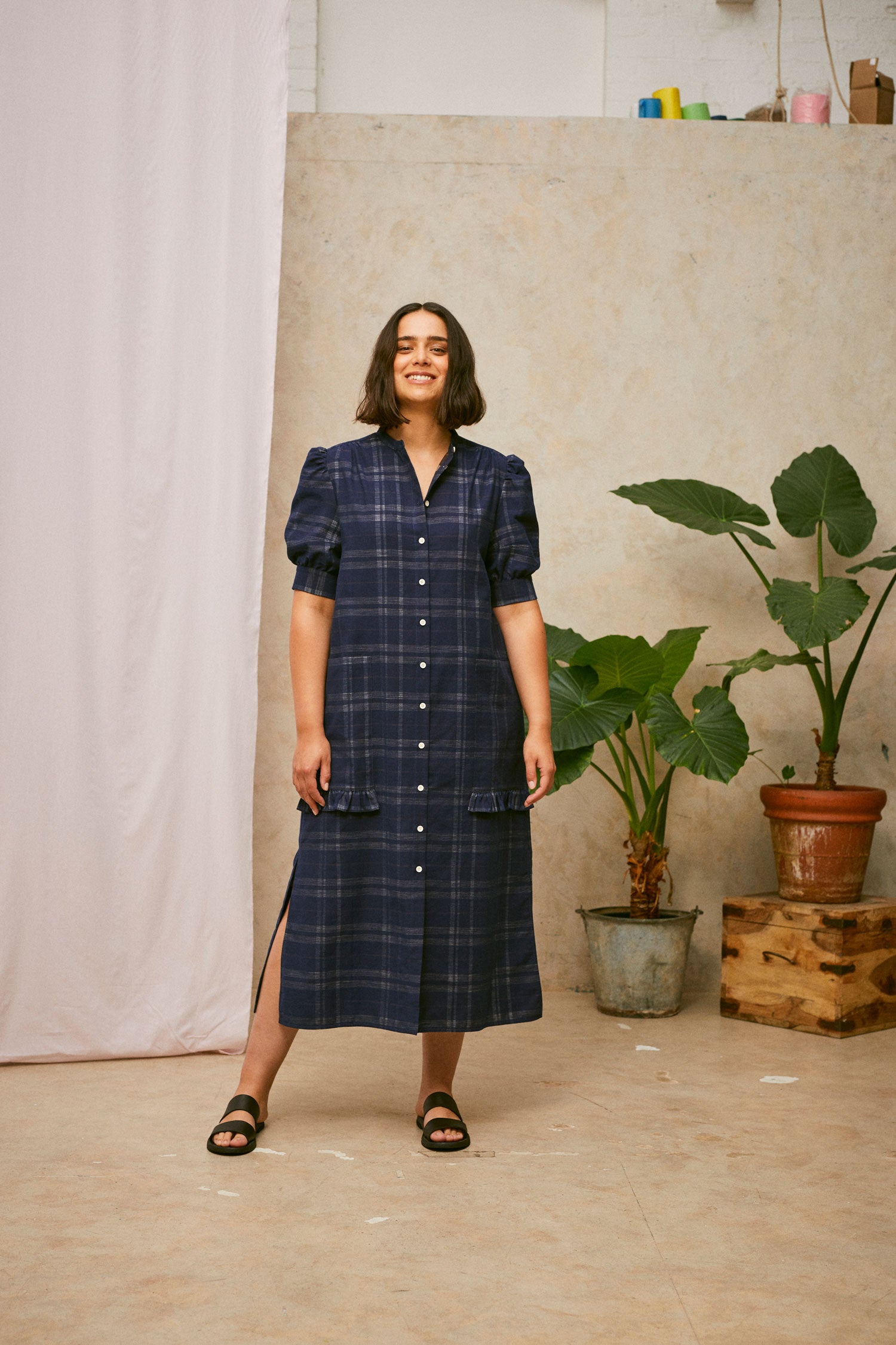 Full length shot of model wearing Saywood's Rosa navy check puff sleeve shirtdress, loosely worn without a belt. The model is smiling. Worn with black sandals. A plant and drop of pink fabric can be seen in the background.