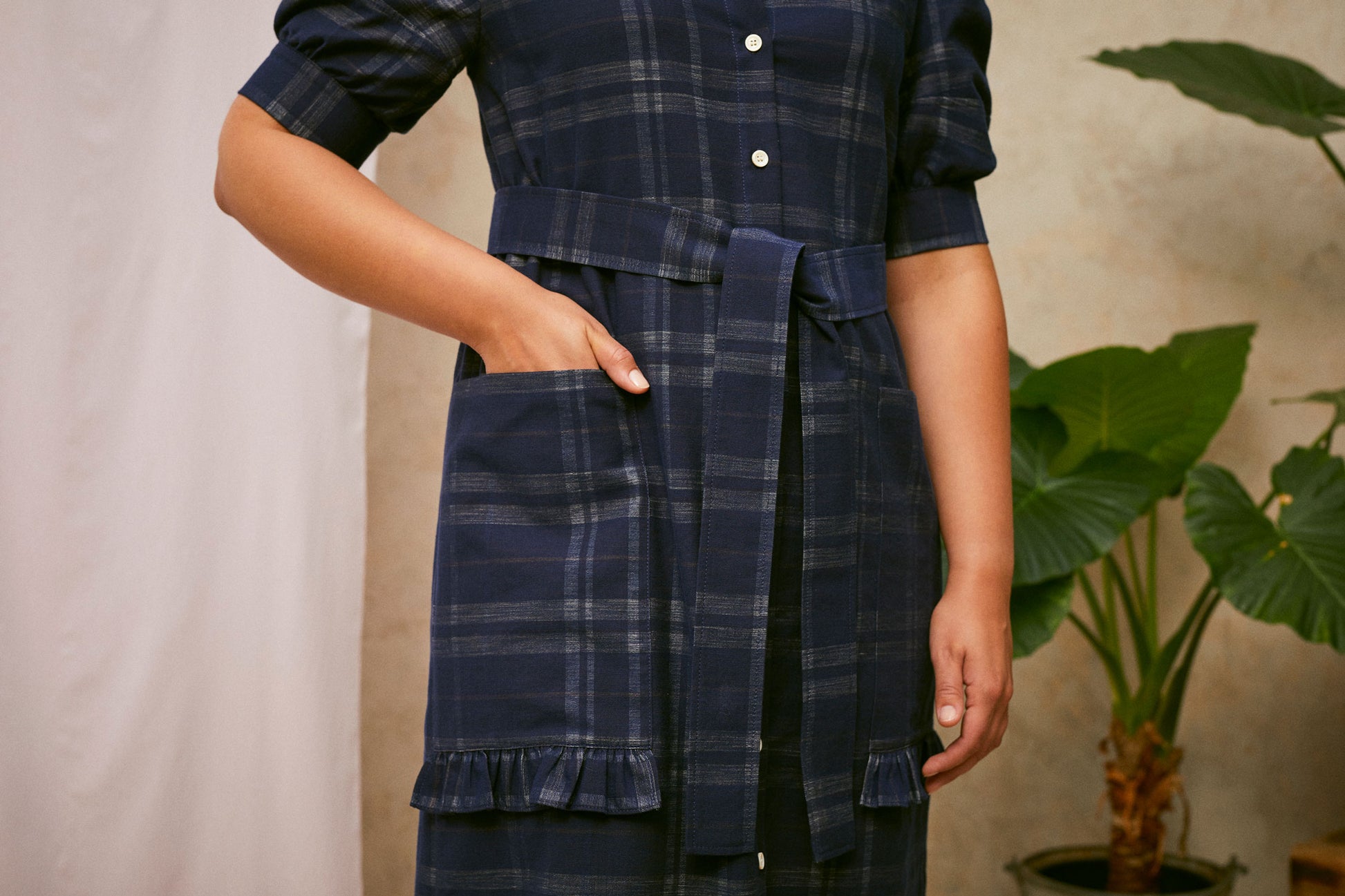Close up of Saywood's Rosa navy check shirtdress, with the belt tied at the waist. Model has one hand in the patch pocket, wthe other hangs to her side. Ruffles at the base of the pockets can be seen. A plant and drop of pink fabric can be seen in the background.
