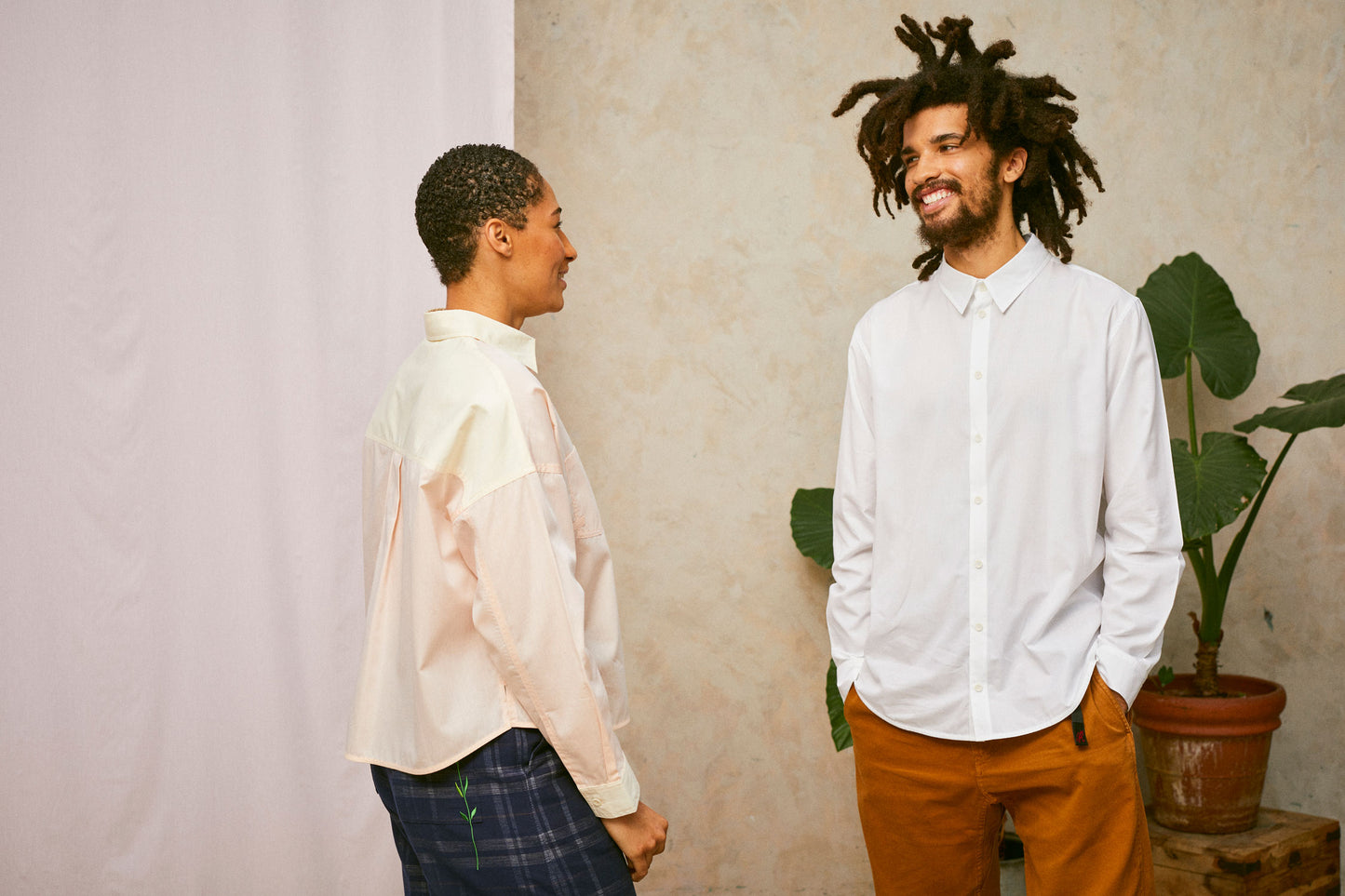 Two models stand laughing and smiling together. The female model has her back angled to the camera, and she wears Saywood's Lela patchwork shirt in pastel orange and yellow. The male model faces her, wearing Saywood's white mens shirt.
