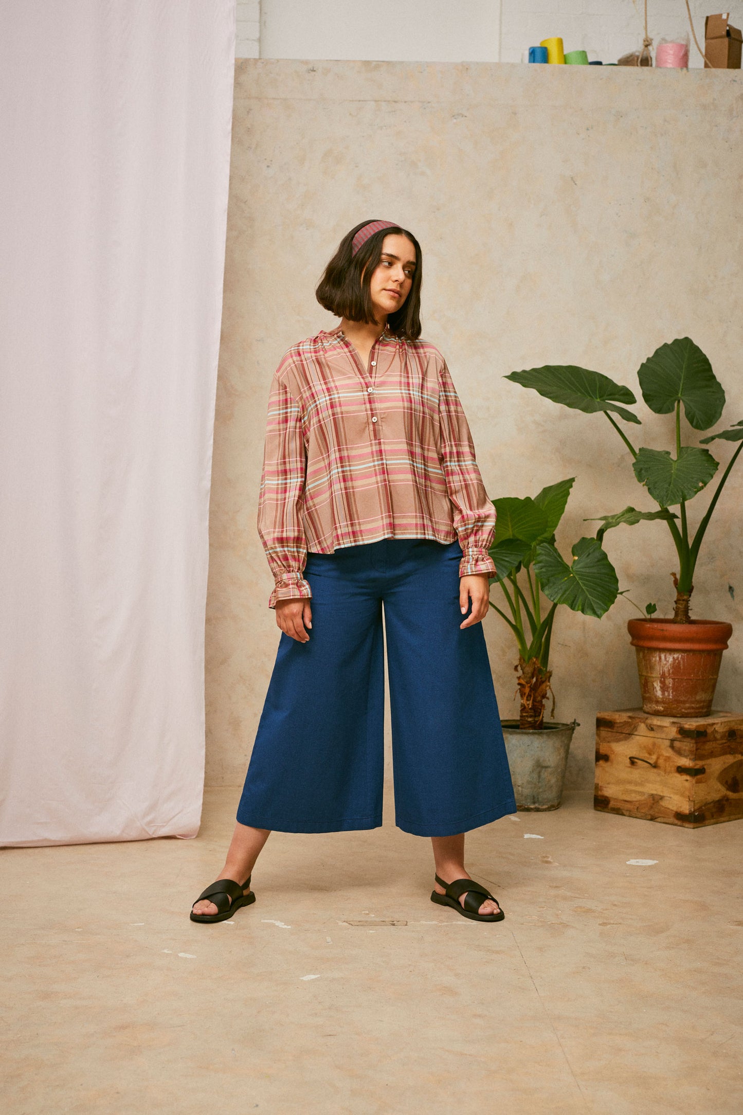 Full length shot of model looking to the side. She wears Saywood's pink check shirt, the Marie A-Line Blouse, with natural indigo Amelia wide leg trousers and black sandals. The red check Heidi Headband is worn in her her. A plant and drop of pink fabric can be seen in the background.
