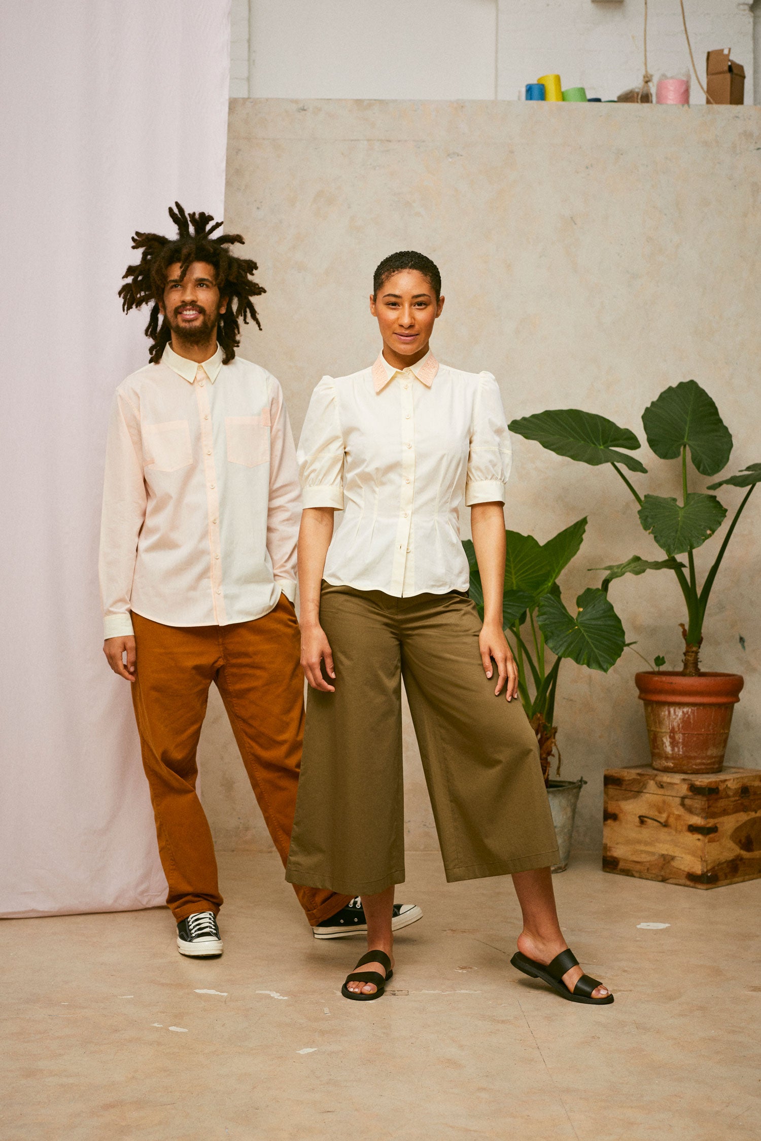 Full length shot of male and female model. He wears Saywood's Eddy Mens mens patchwork Shirt with patch pockets in orange and yellow. Worn with tabacco trousers and black Converse. She wears Saywood's Joni yellow blouse with orange lace collar and Amelia khaki wide leg trousers and black sandals. A plant and drop of pink fabric can be seen in the background.
