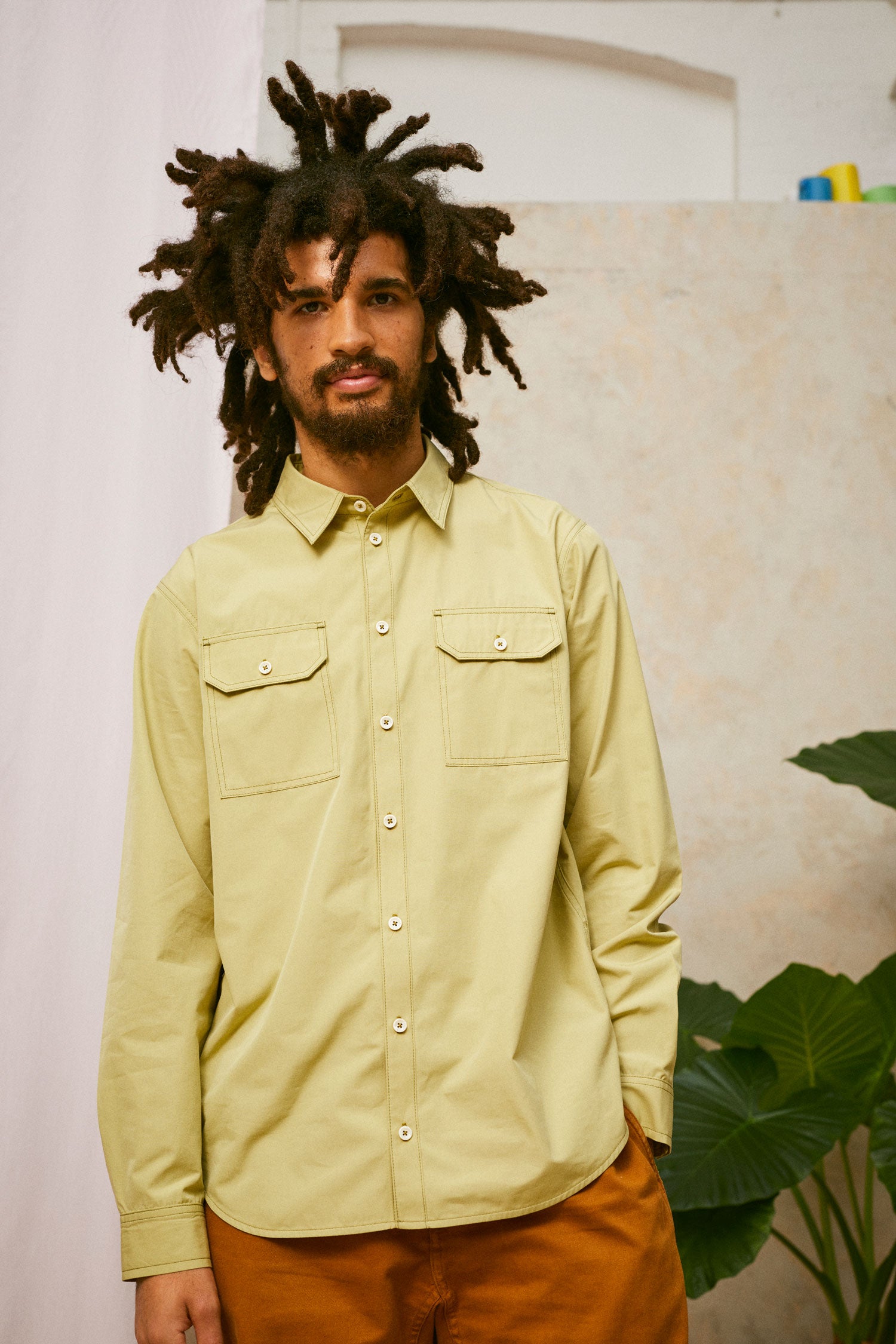 Close up of model wearing Saywood's Eddy Mens Khaki Shirt with utility pockets. Worn with tabacco trousers just visible. Model has one hand in his trouser pocket and a plant and drop of pink fabric can be seen in the background.