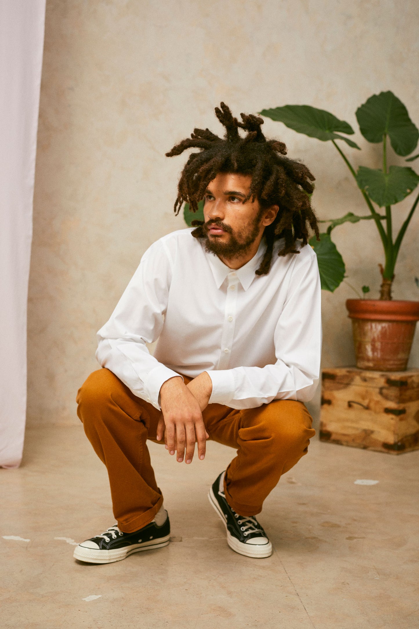 Model is crouching on the floor, with his arms resting on his knee and hands close together. He is wearing Saywood's Eddy Mens mens white Shirt. Worn with tabacco trousers and black Converse. A plant and drop of pink fabric can be seen in the background.