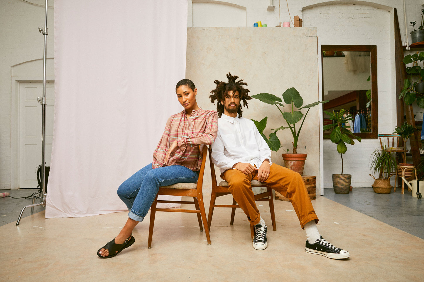 Image shows a males and female model sitting on chairs with their back together, each with one leg out. She wears Saywood's pink check shirt, the Marie A-Line Blouse, with jeans and black sandals. He wears Saywood's Eddy mens white shirt with patch pockets, tobacco trousers and black Converse. A pink drop of fabric and plants are seen in the background.