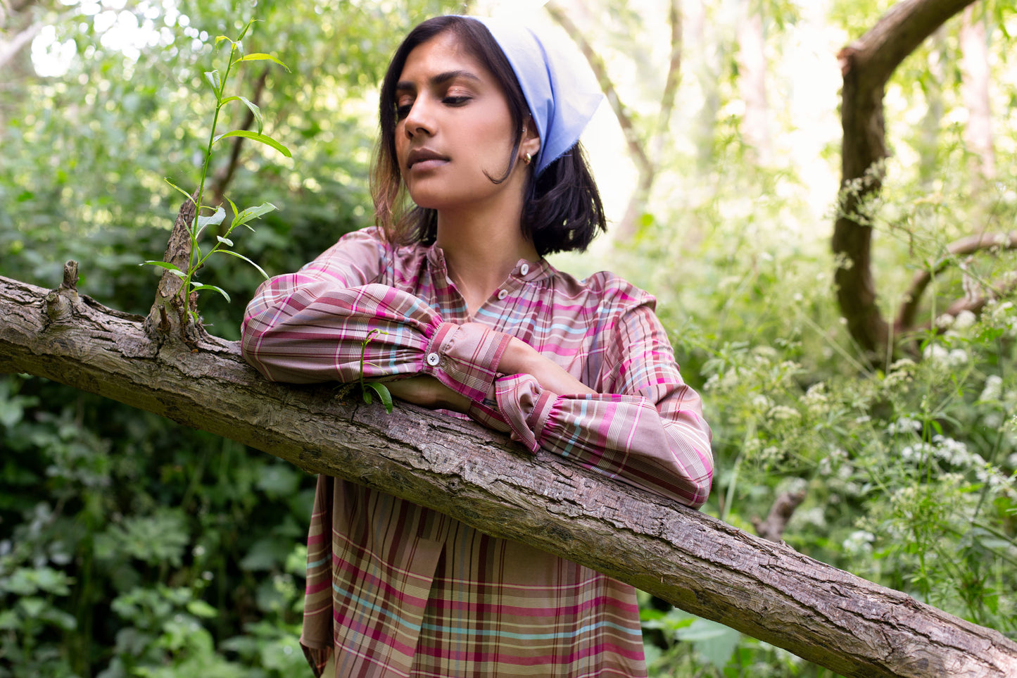 Model leans on a angled tree brand, surrounded by greenery. She wears Saywood's pink check shirt, the Marie A-Line Blouse, with a pale blue recycled cotton bandana in her hair.