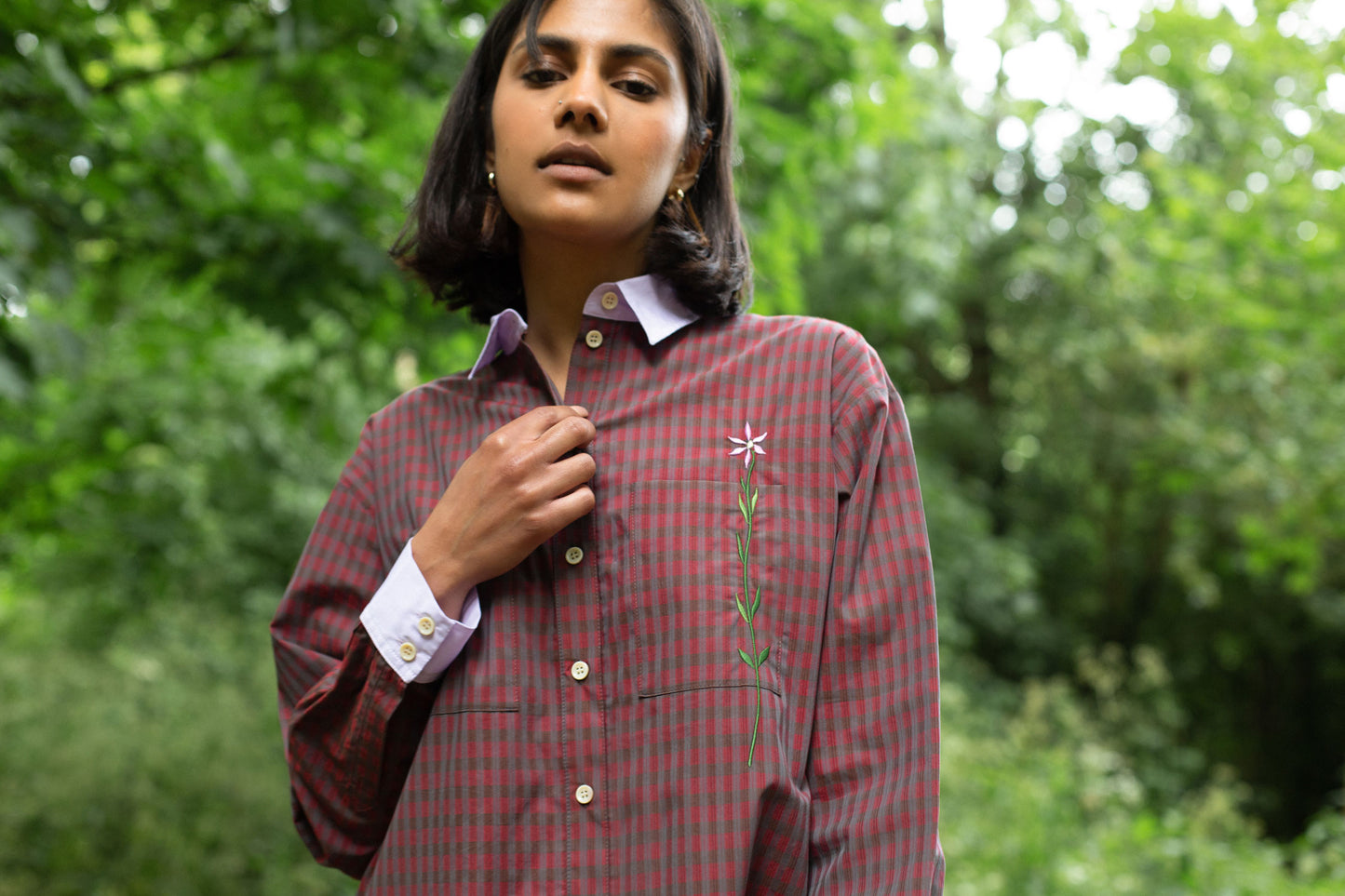 Model wears the Saywood Etta Oversized Red Check Shirtdress in sustainably sourced deadstock cotton. She stands surrounded by trees. The signature flower embroidery can be seen on the chest pocket, and the contrast lilac collar and cuffs.
