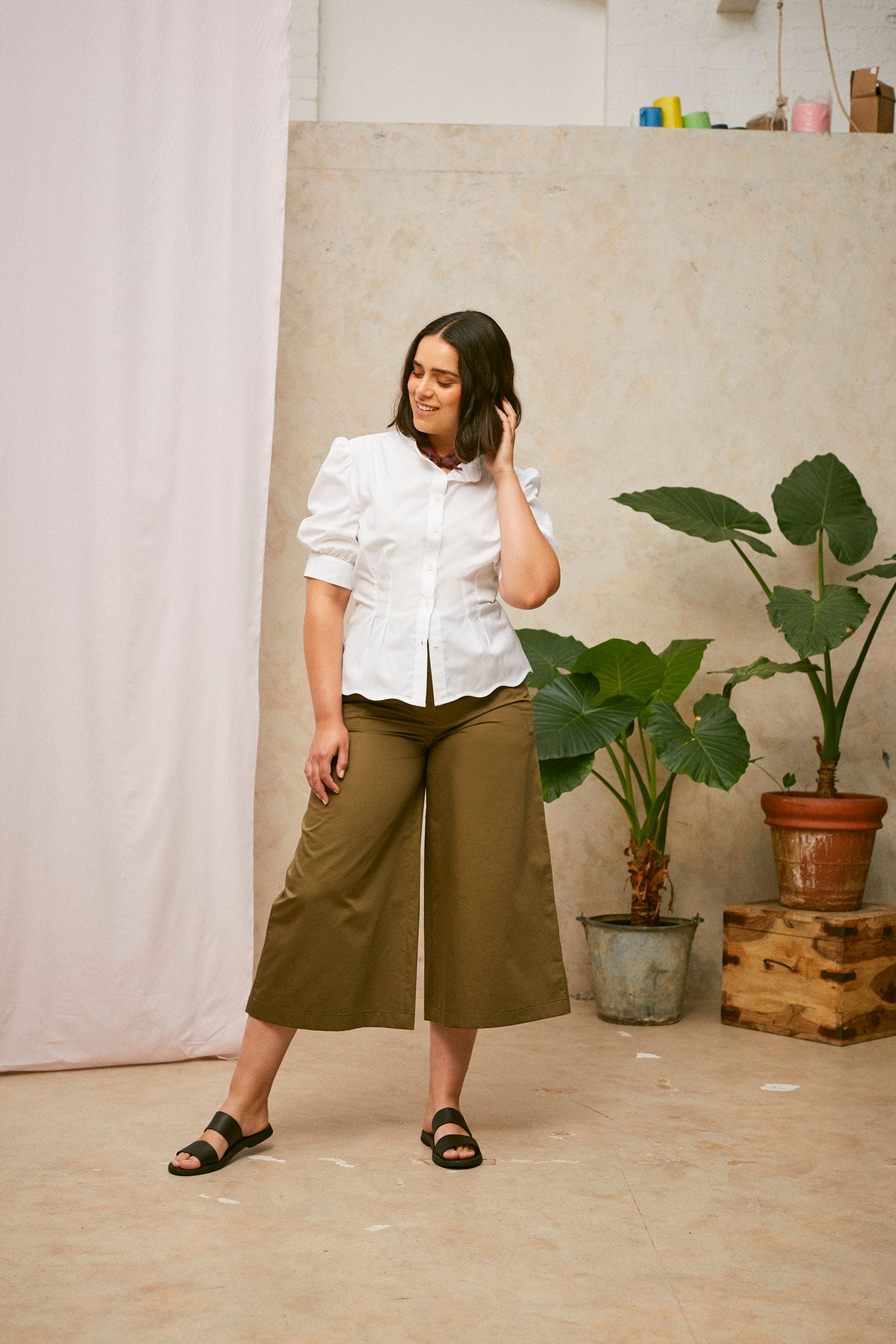 Model stands smiling, wearing Saywood's Joni puff sleeve blouse in white with scalloped hem, worn with khaki Amelia wide leg trousers and black sandals. A plant and drop of pink fabric can be seen in the background. 