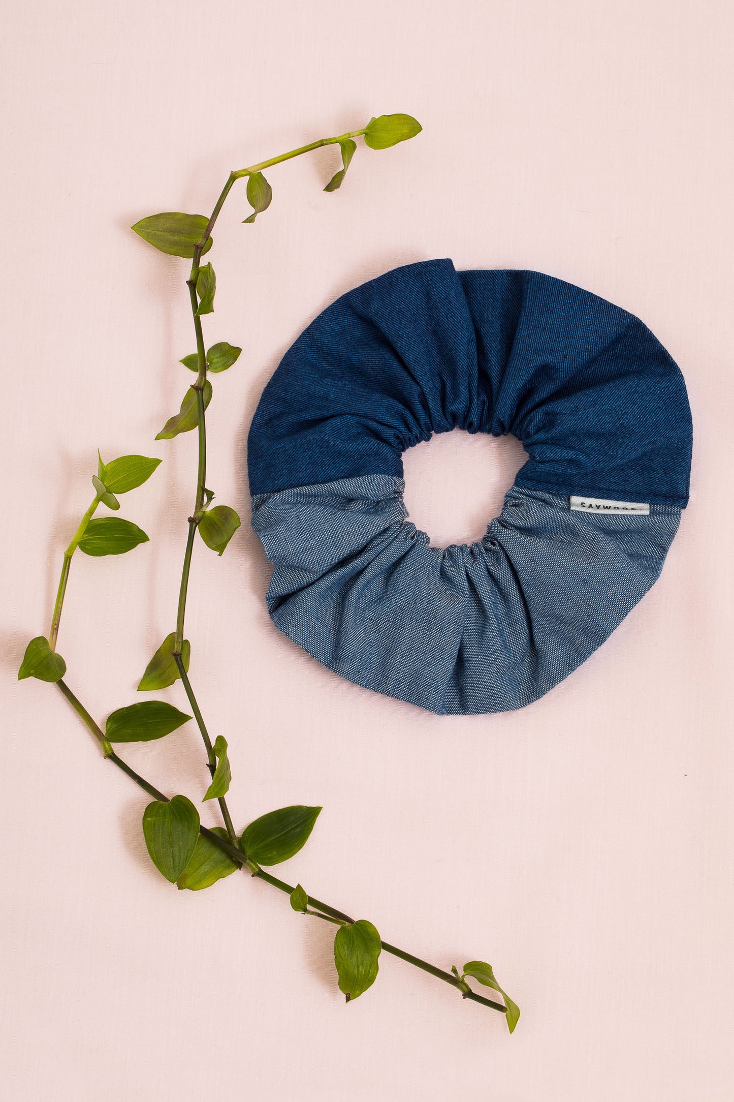 Patchwork scrunchie, made in mid wash and light wash Japanese denim. A Saywood label is stitched into the seam. The scrunchie sits on a pale pink backdrop with green foliage.