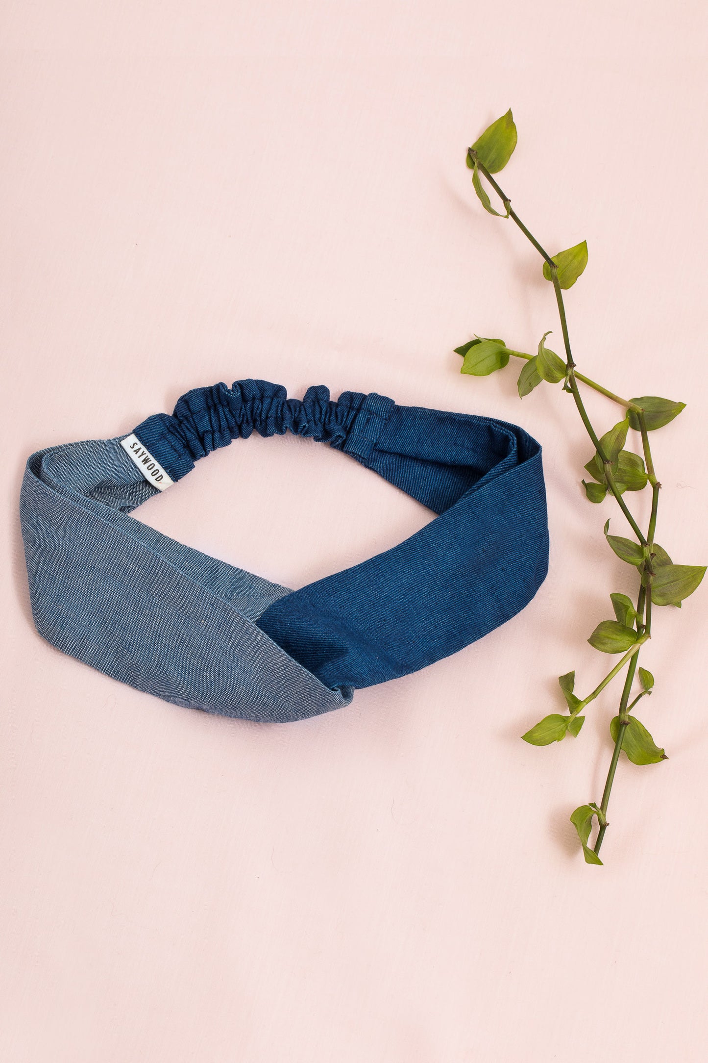 Saywood's Thandi Headband with twist effect, made from contrasting mid wash and light wash natural indigo Japanese denim. Laid on a pale pink background with green foliage.