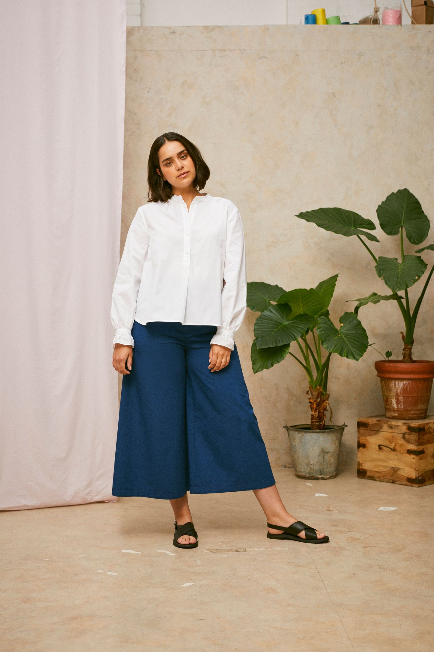 Full length shot of model wearing Saywood's Amelia denim trousers, wide leg culotte shape in Japanese denim, with black sandals. Worn with the white blouse, Marie a-line-shirt. A plant and drop of pink fabric can be seen in the background.