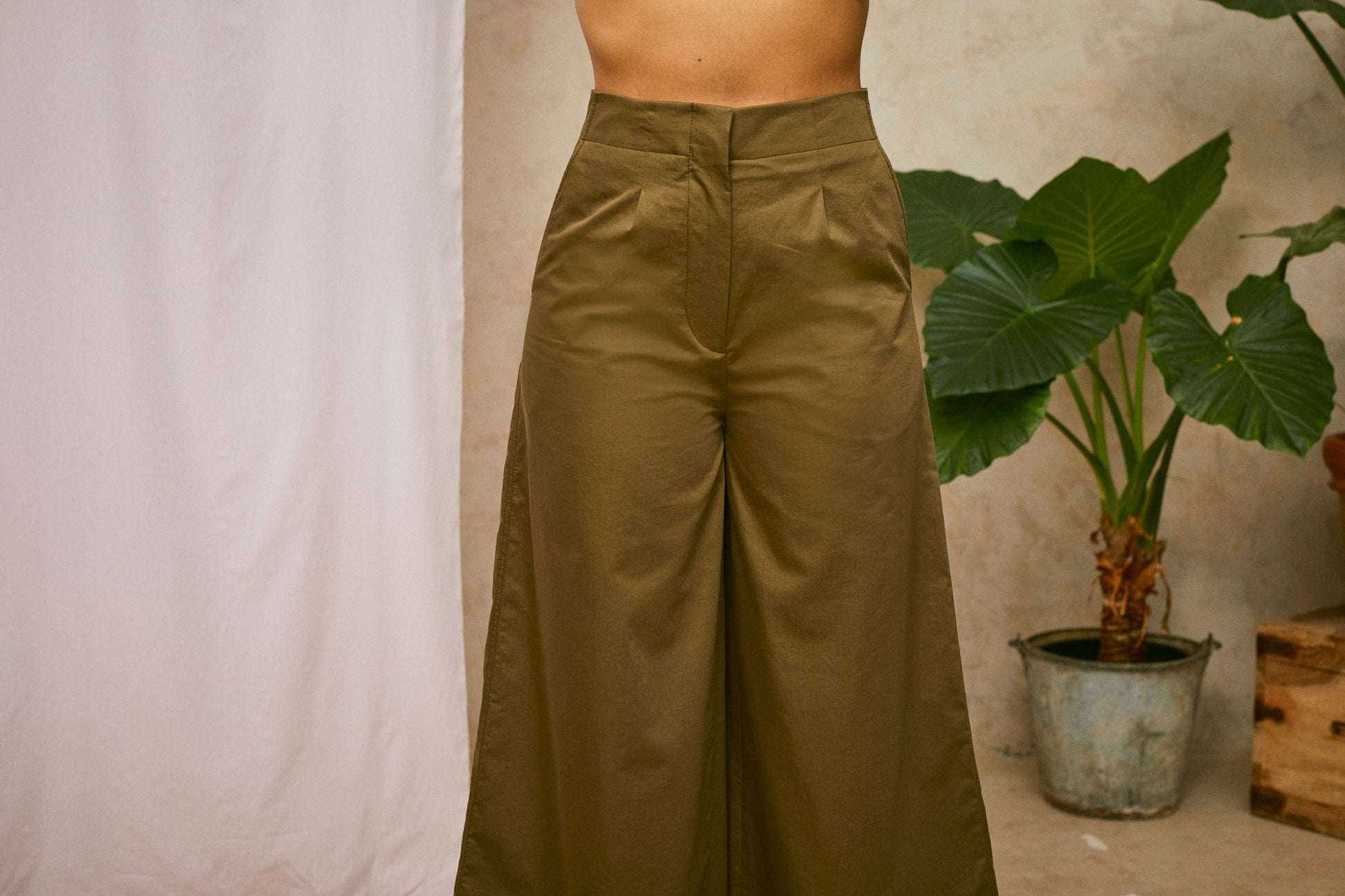 Cropped image of models wearing Saywood's Amelia khaki wide leg trouser, front waist view. A plant and drop of pink fabric can be seen in the background.