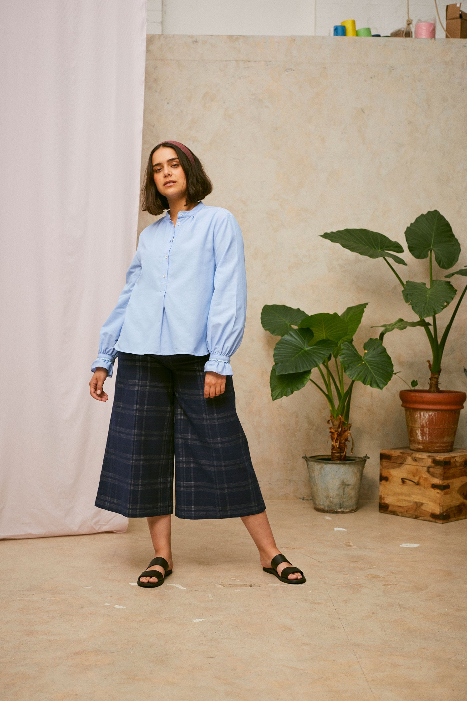 Full length shot of model wearing Saywood's Amelia check trousers, wide leg culotte shape in navy check, with black sandals. Worn with the pale blue shirt, the Marie a-line blouse. A plant and drop of pink fabric can be seen in the background.