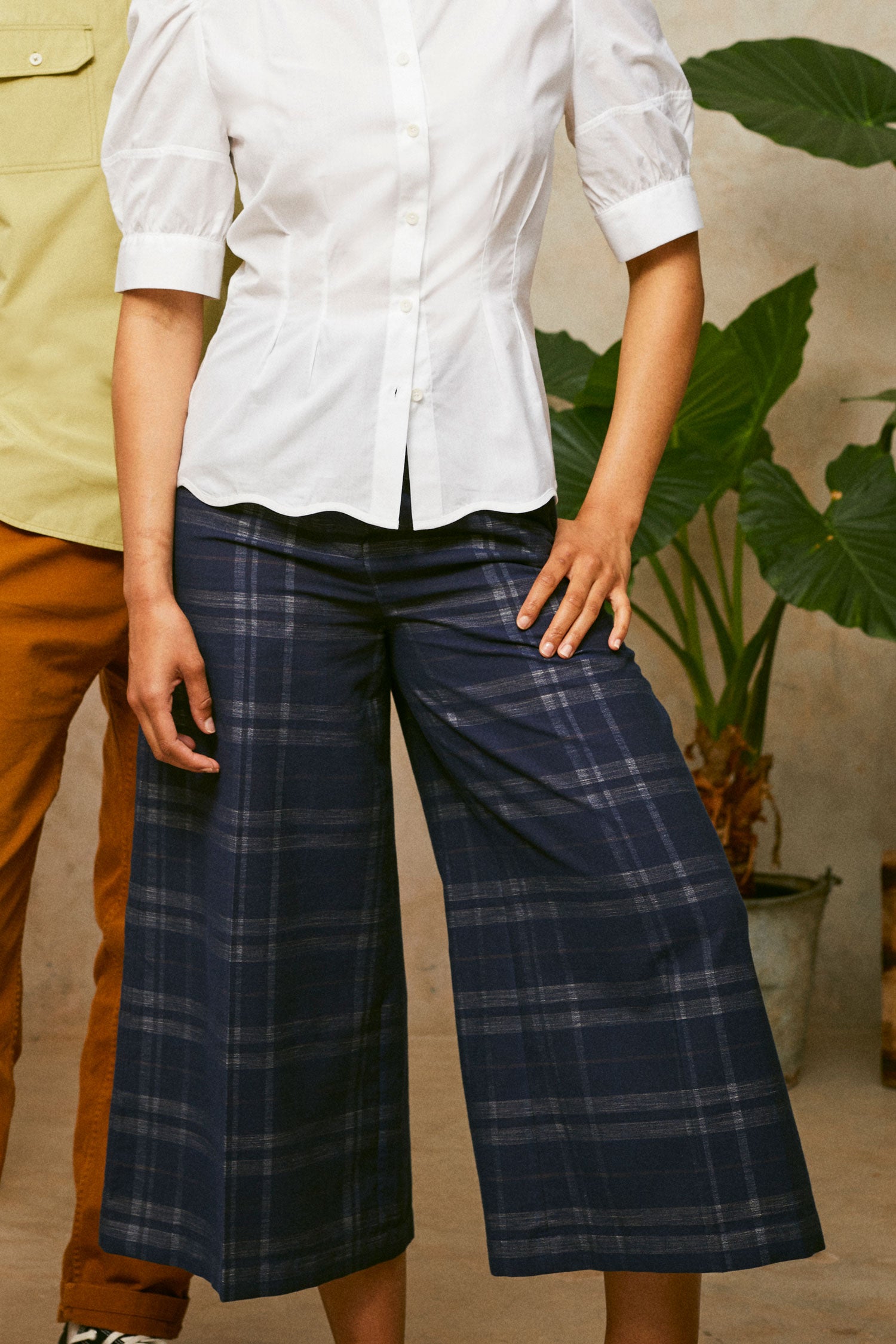 Close up of model wearing Saywood's Amelia check trousers, wide leg culotte shape in navy check, with black sandals. Worn with the white puff sleeve shirt, Joni blouse. A plant and another model can be seen in the background.
