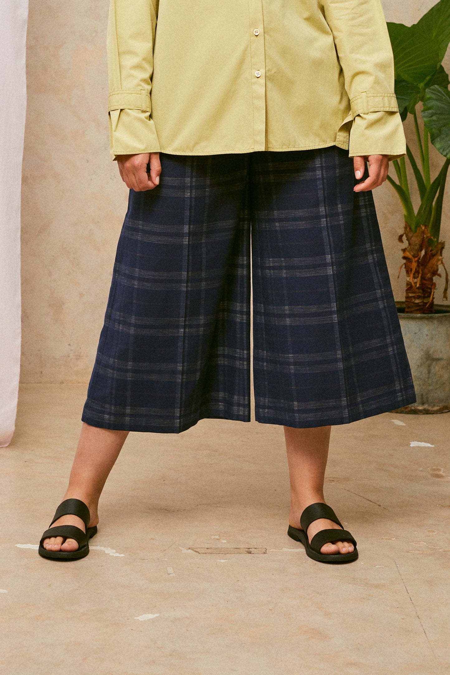 Close up of model wearing Saywood's Amelia check trousers, wide leg culotte shape in navy check, with black sandals. Worn with the olive shirt, Zadie boyfriend shirt. A plant and drop of pink fabric can be seen in the background.