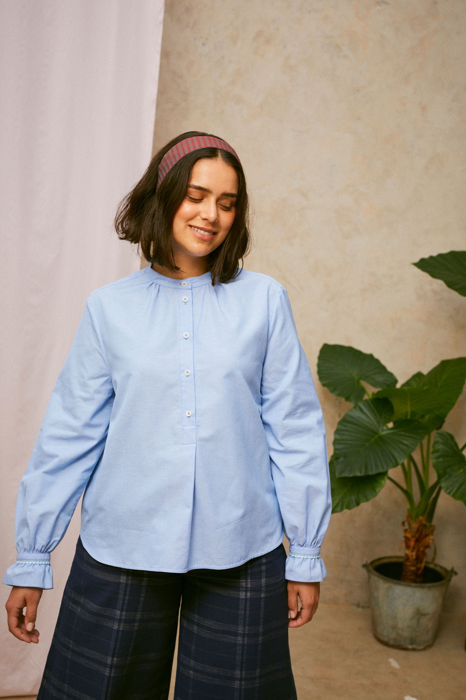 Model wears Saywood's pale blue shirt, the Marie A-Line Blouse, with navy check Amelia wide leg trousers. The red check Heidi Headband is worn in her her. A plant and drop of pink fabric can be seen in the background. 