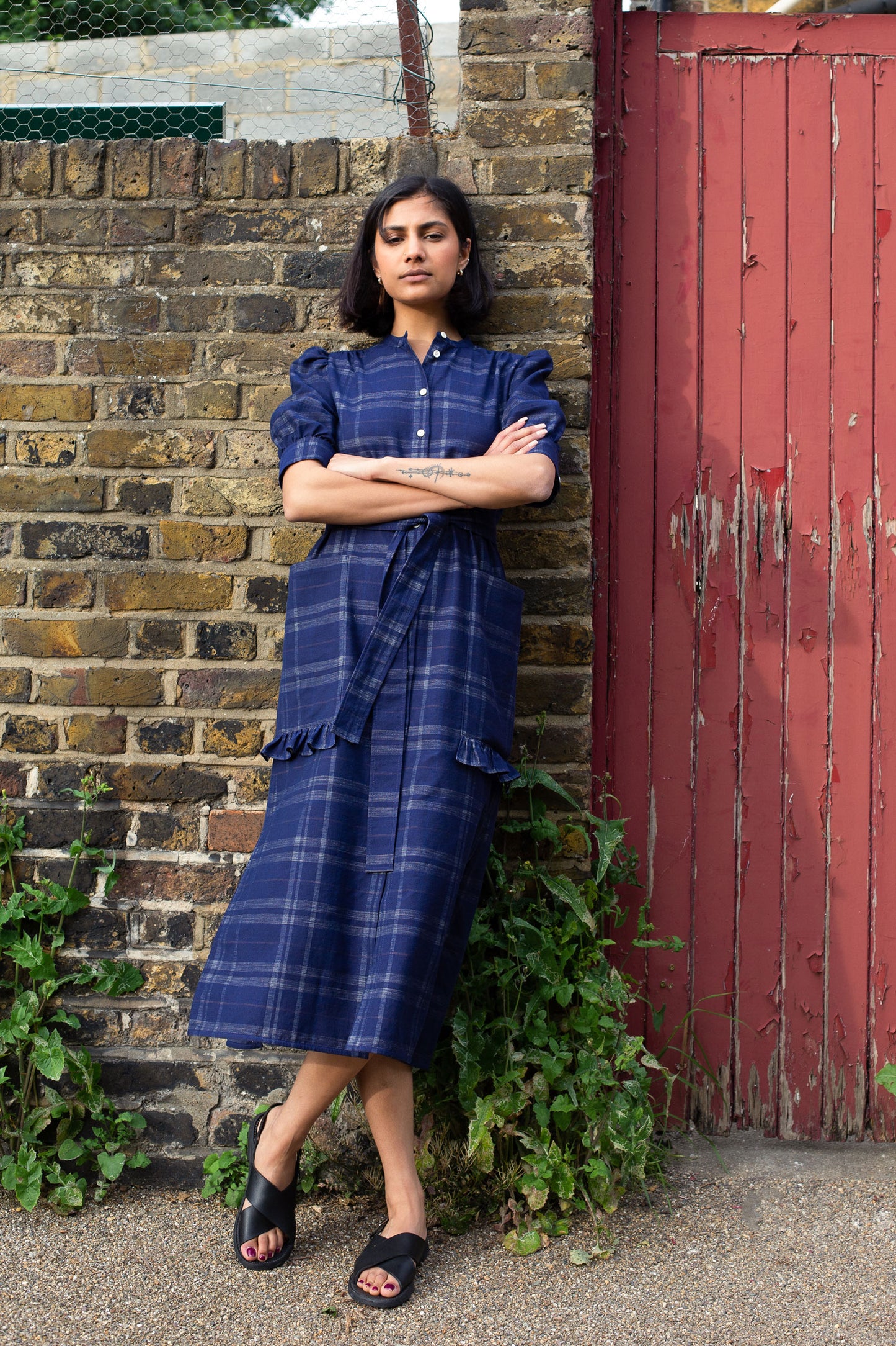 Model leans against a brick wall with a red gate door next to her. She has her arms folded and wears Saywood's Rosa puff sleeve dress in navy check with black sandals. Plants can be seen growing out near her feet.