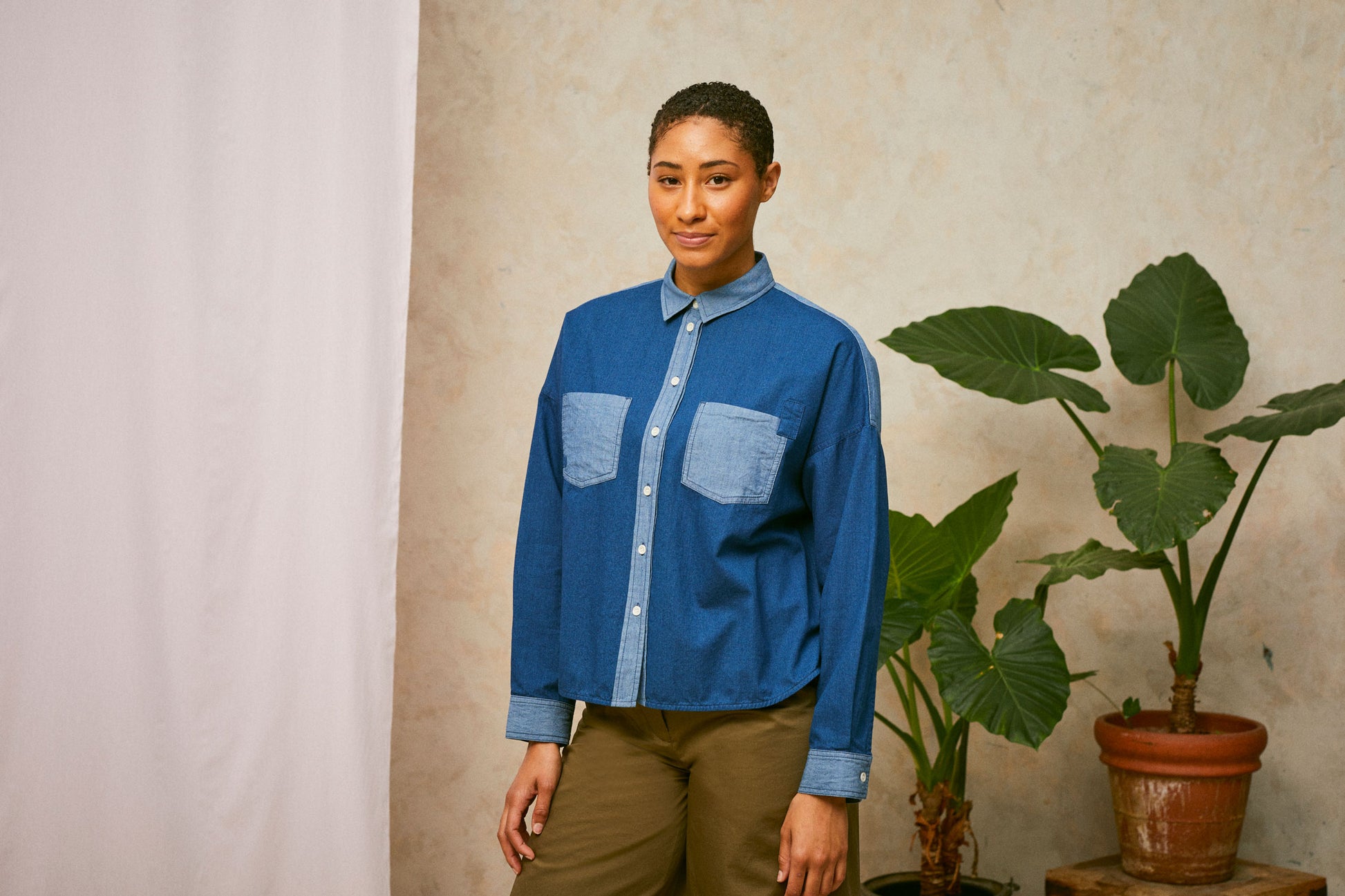Model stands in front of a pink drop of fabric and two plants in the background. She wears Saywood's Japanese Denim Lela Patchwork Shirt, with light wash contrast cuffs, pockets and collar. Worn with khaki Amelia Trousers.