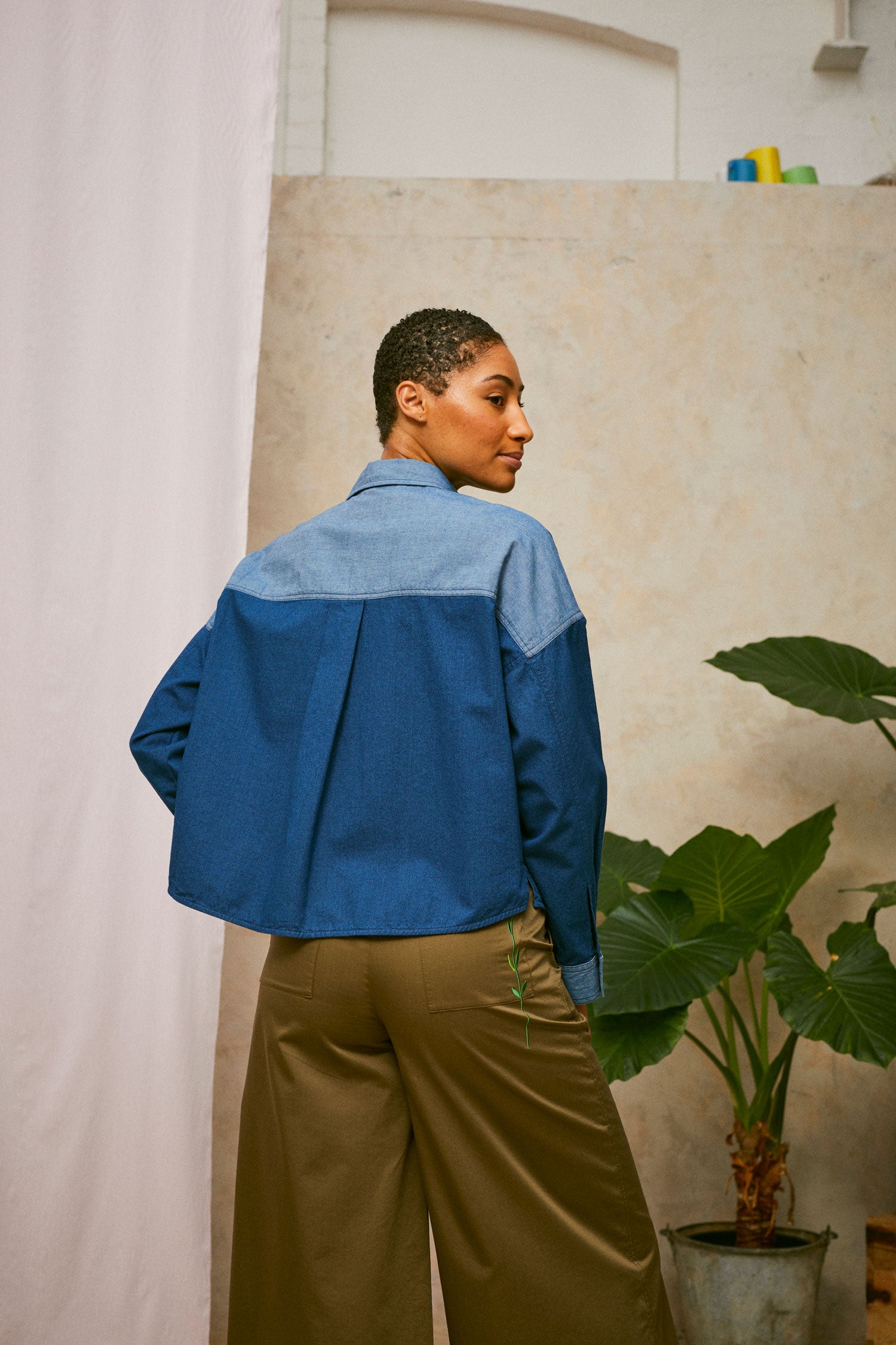 Model stands in front of a pink drop of fabric and two plants in the background. She wears Saywood's Japanese Denim Lela Patchwork Shirt, with light wash contrast back neck yoke and cuffs. Worn with khaki Amelia Trousers. Back view