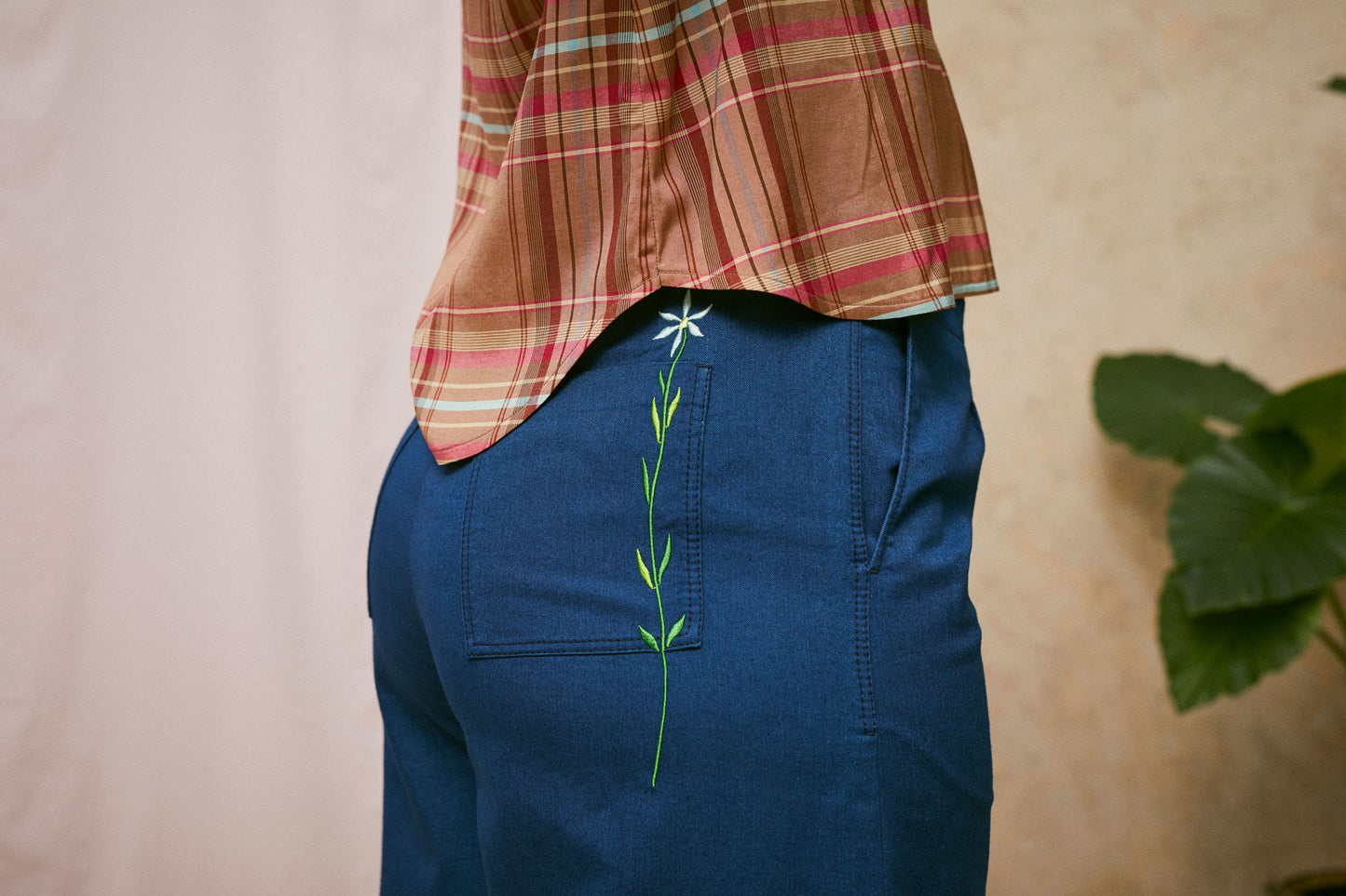 Close up side view of model wearing Saywood's Amelia denim trousers, wide leg culotte shape in Japanese denim. The blue flower embroidery can be seen on the back patch pocket. Worn with the pink check blouse, Marie a-line-shirt. A plant and drop of pink fabric can be seen in the background.