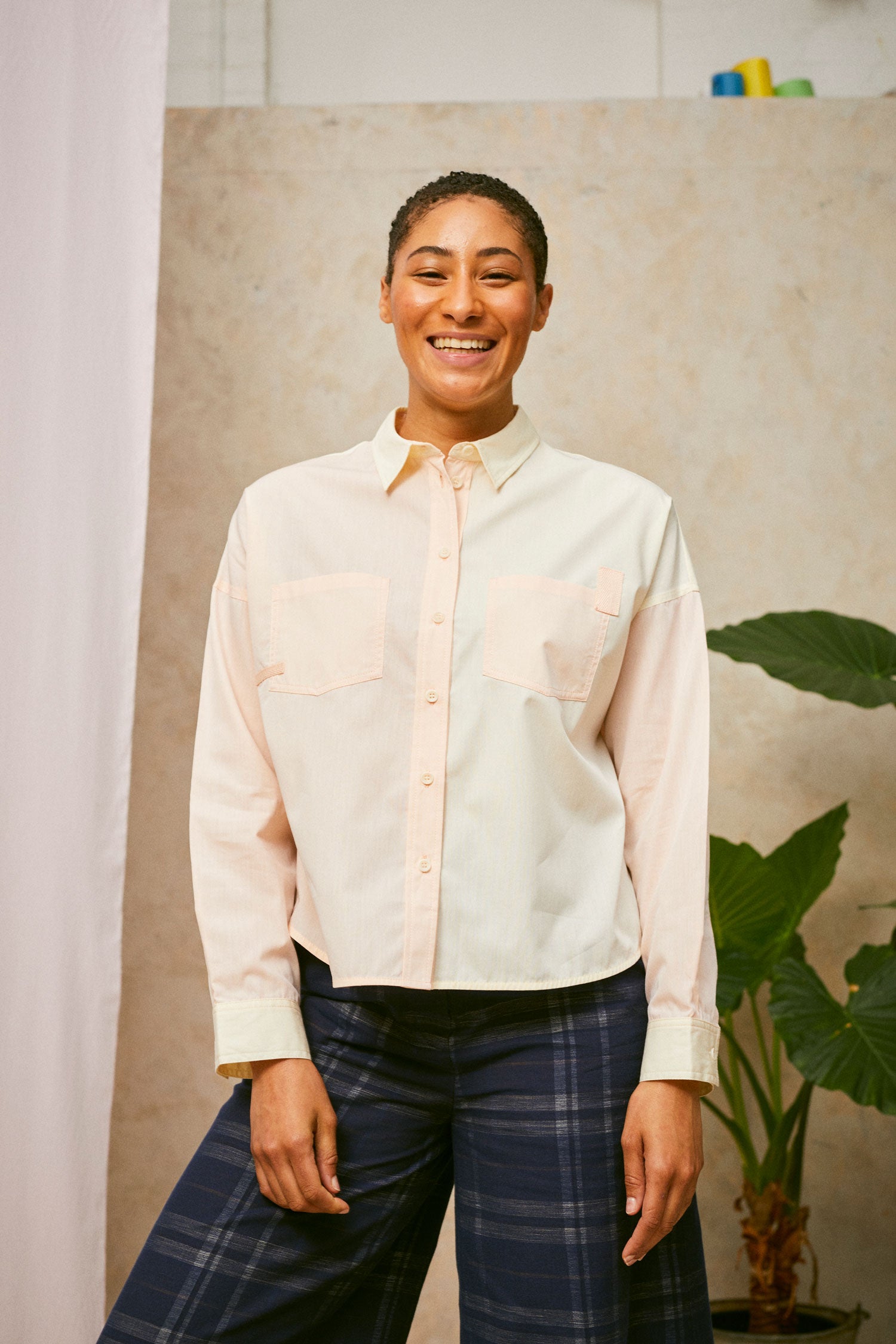 Cropped upper body image of model smiling, wearing the Saywood Lela patchworkshirt in pastel orange and yellow, with patch pockets on the chest. Worn with navy check trousers. A plant can be seen in the background.