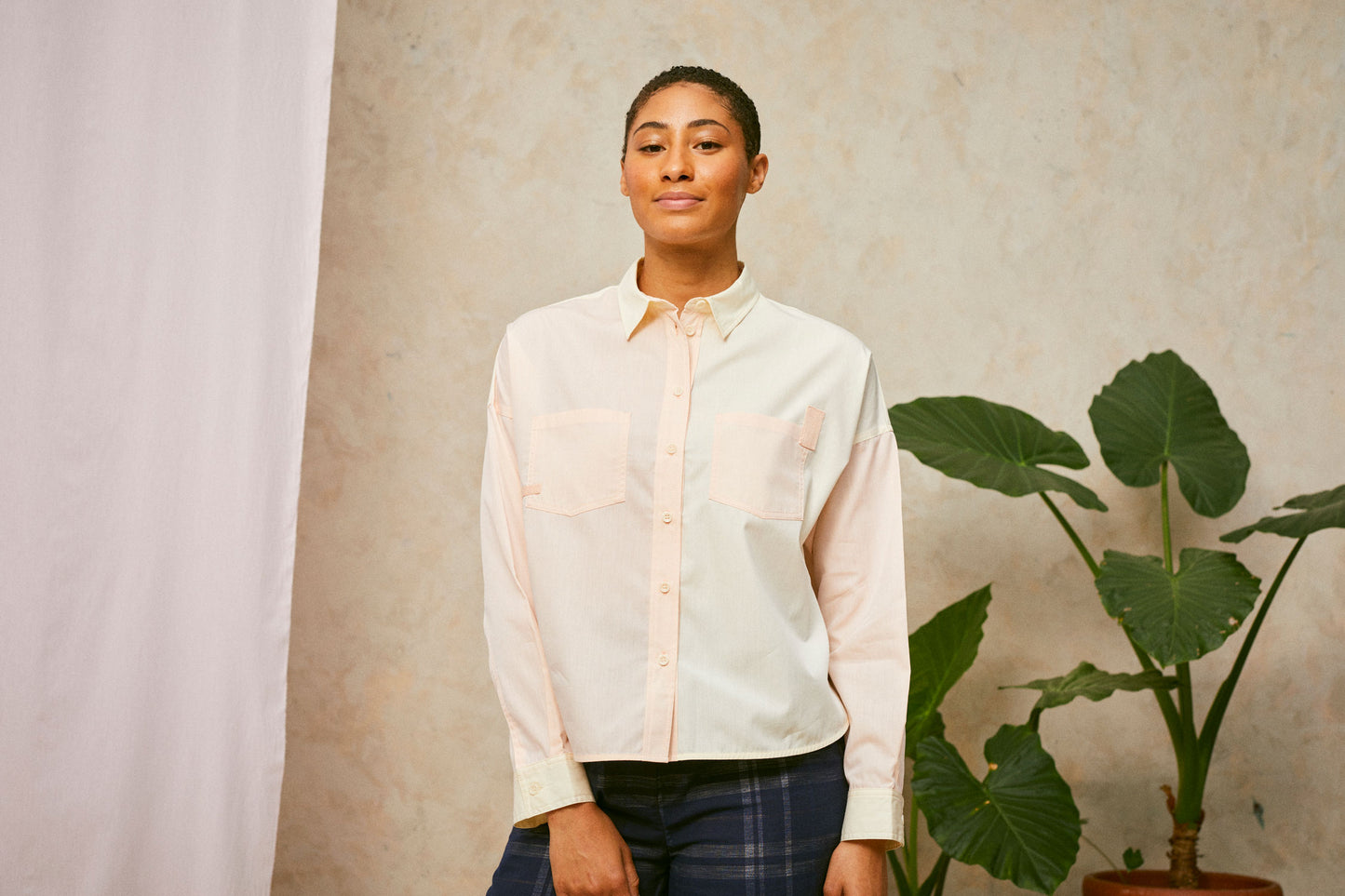 Model stands front on facing the camera, wearing Saywood's Lela pastel orange and yellow patchwork shirt, with navy check trousers. A plant and pink fabric can be seen in the background.