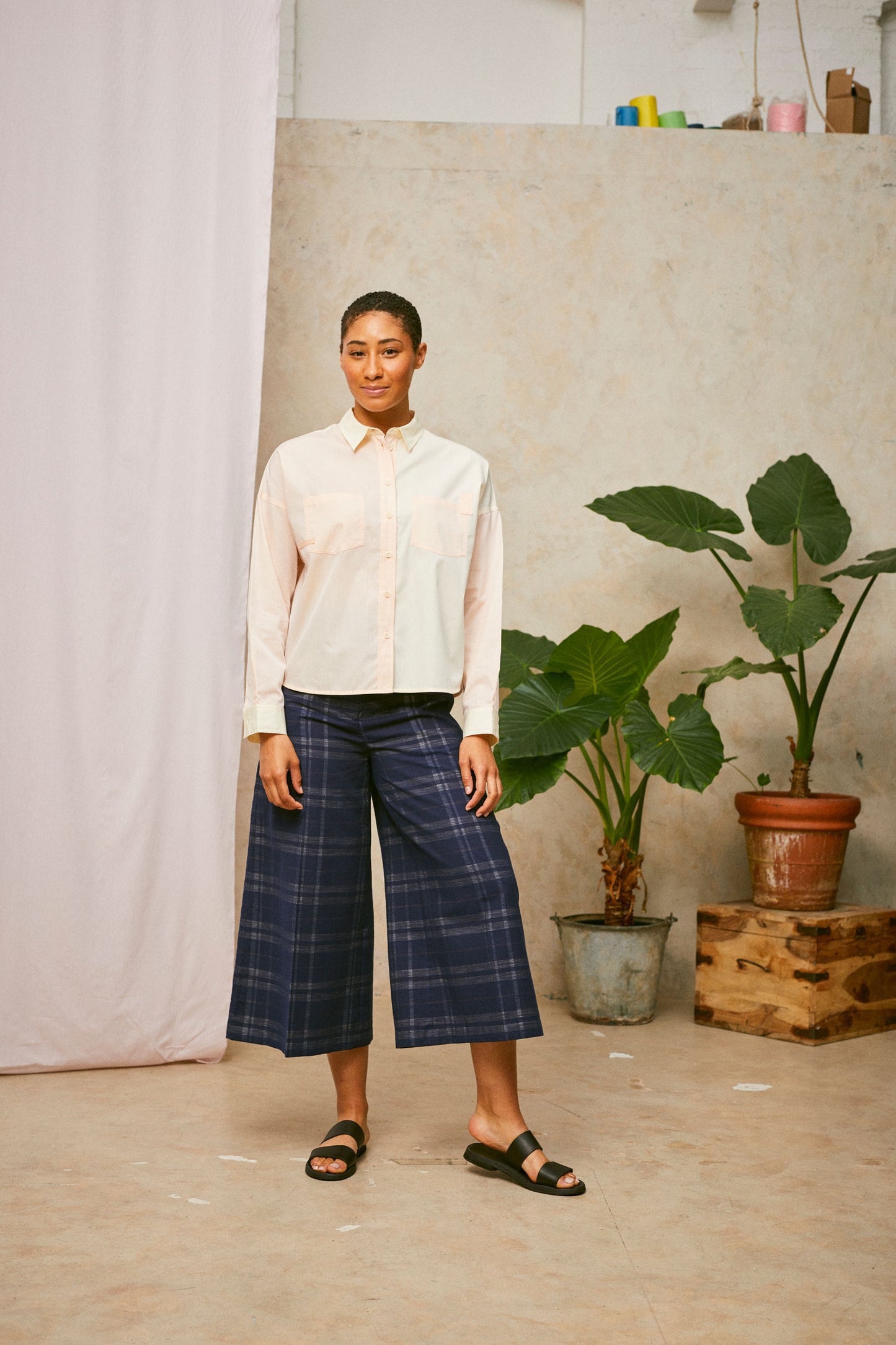 Model stands, with several plants in the background and pink fabric hanging on the left. She wears the Lela Patchwork Shirt in pastel orange and yellow, worn with navy check trousers and black sandals.