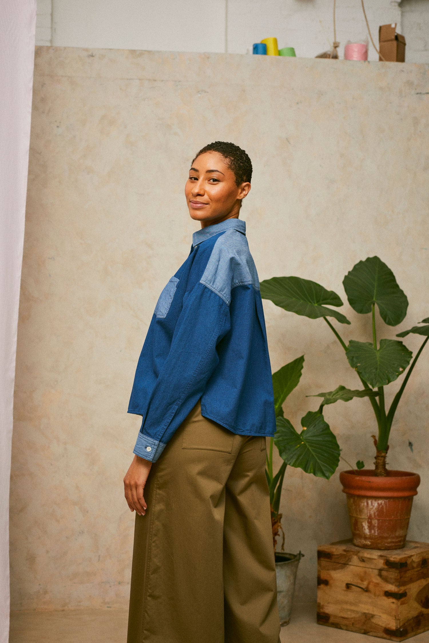 Model stands in front of a pink drop of fabric and two plants in the background. She wears Saywood's Japanese Denim Lela Patchwork Shirt, with light wash contrast cuffs, pockets, collar and yoke. Worn with khaki Amelia Trousers. Side view