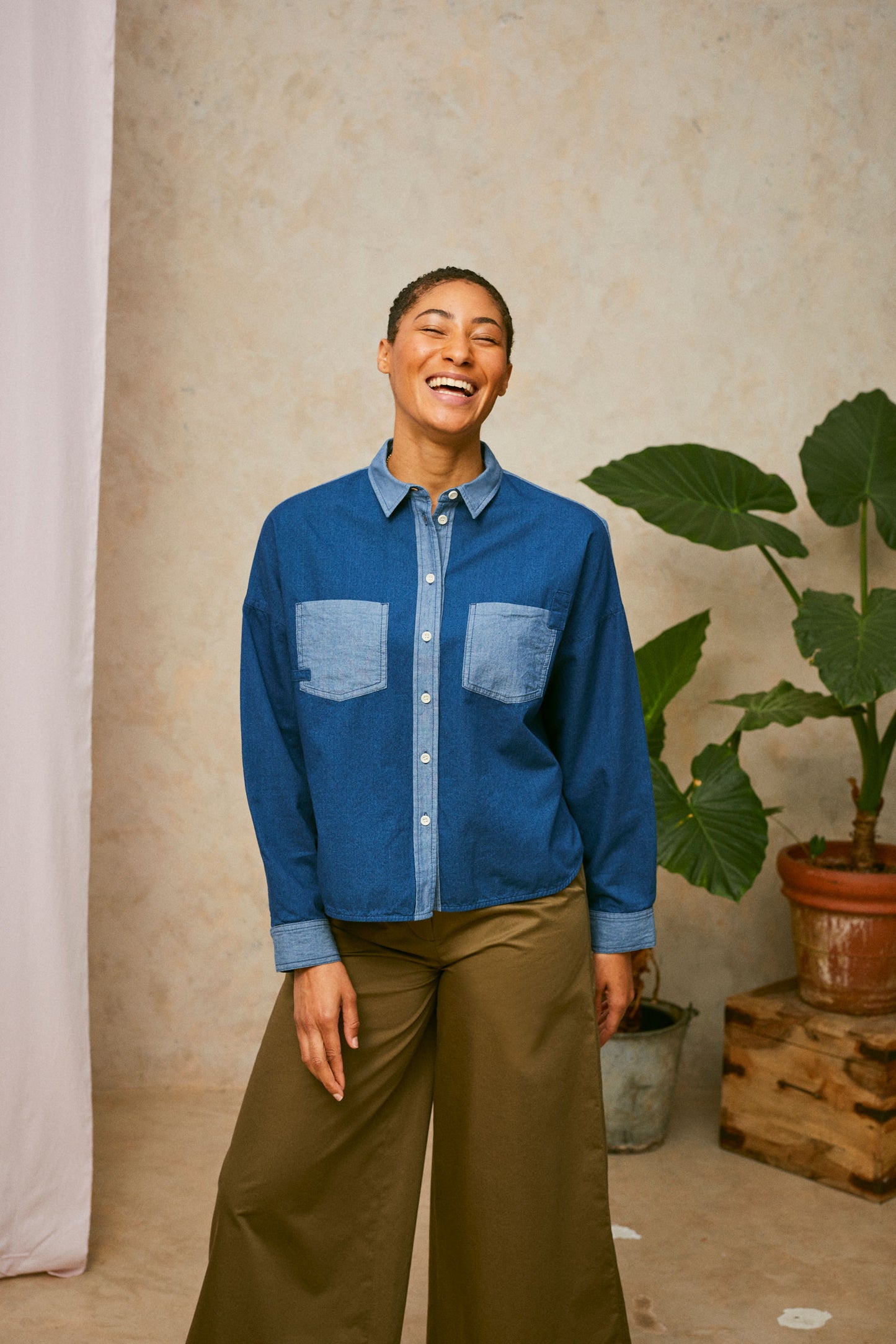 Model stands in front of a pink drop of fabric and two plants in the background. She wears Saywood's Japanese Denim Lela Patchwork Shirt, with light wash contrast cuffs, pockets and collar. Worn with khaki Amelia Trousers.