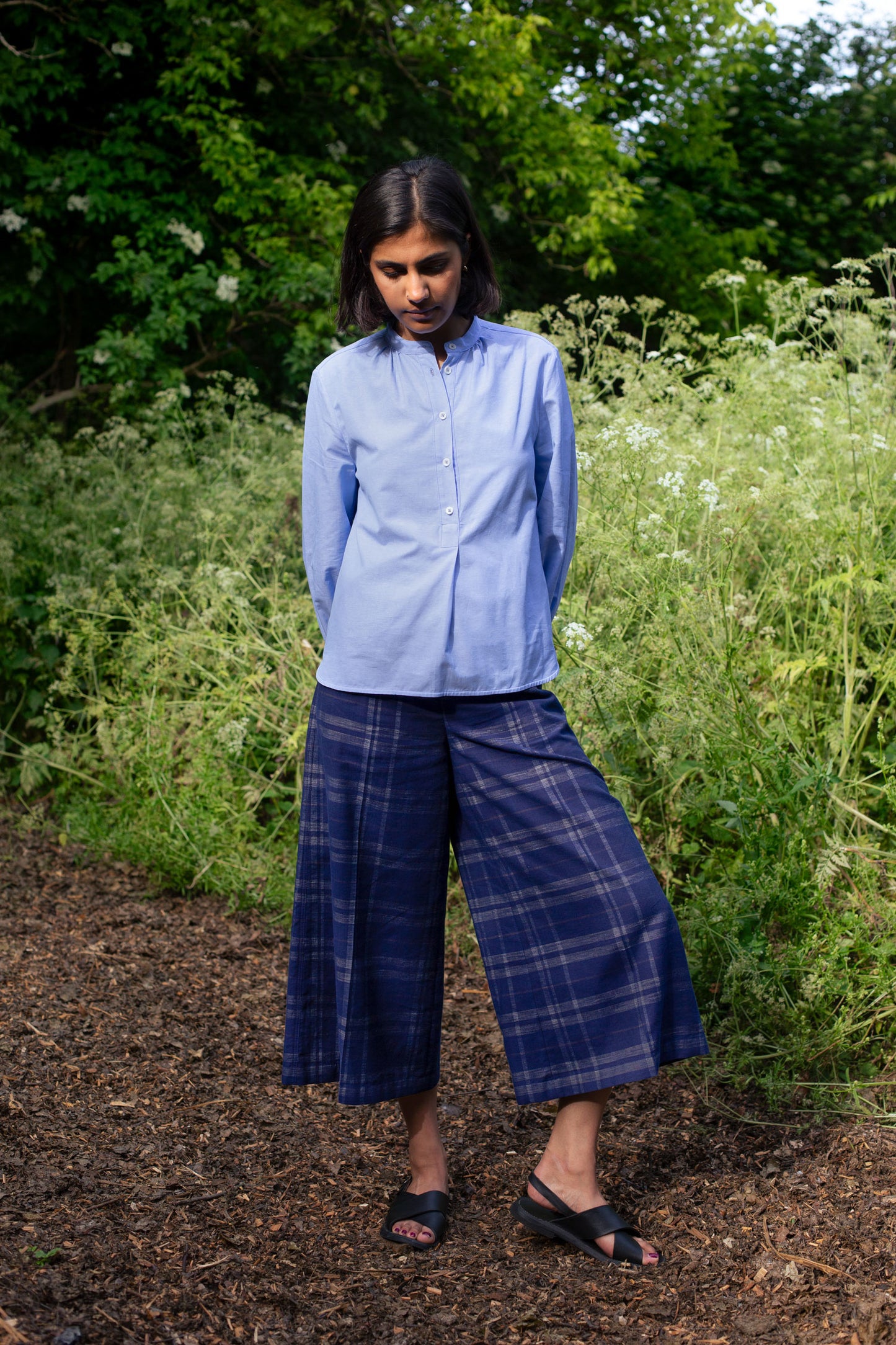 Full length shot of model wearing Saywood's Amelia check trousers, wide leg culotte shape in navy check, with black sandals. Worn with the pale blue shirt, the Marie a-line blouse. She is standing outside surrounded by wild plants and greenery.