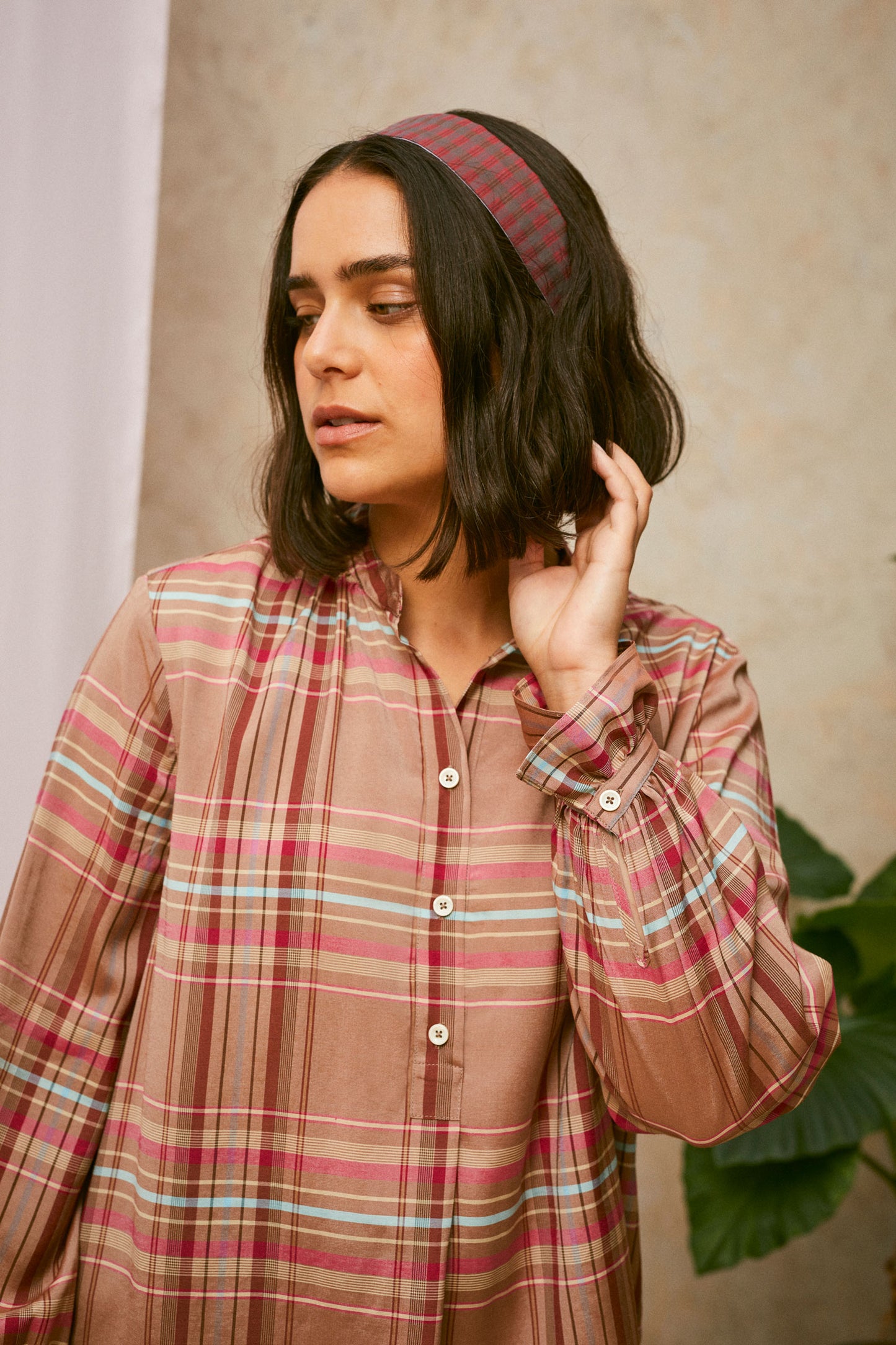 Model stands, with one hand to her head. She wears Saywood's pink check shirt, with the red check Heidi Headband worn in her her. A plant and drop of pink fabric can be seen in the background. 