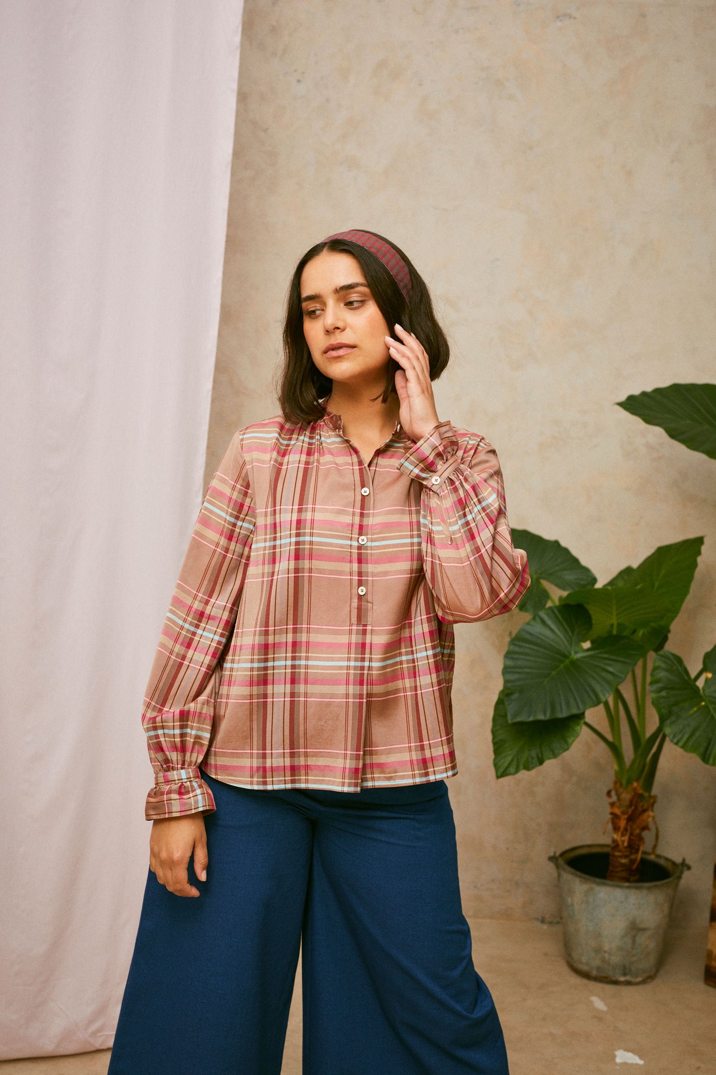Model stands, with one hand to her head. She wears Saywood's pink check shirt, the Marie A-Line Blouse, with natural indigo Amelia wide leg trousers. The red check Heidi Headband is worn in her her. A plant and drop of pink fabric can be seen in the background. 