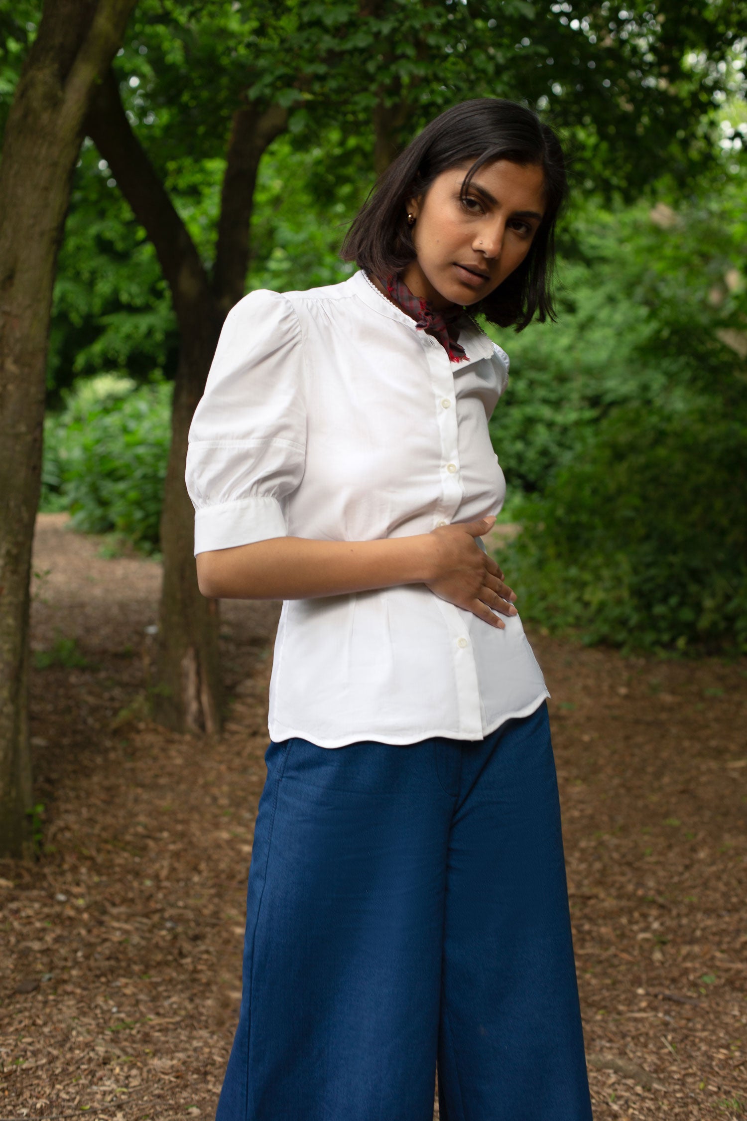 Model stands surrounded by greenery, with one arm across her waist. She wears Saywood's Joni puff sleeve blouse in white with scalloped hem, worn with a red check neckerchief tied round her neck. On the bottom she wears Saywood's Amelia wide leg trousers in natural indigo Japanese denim.