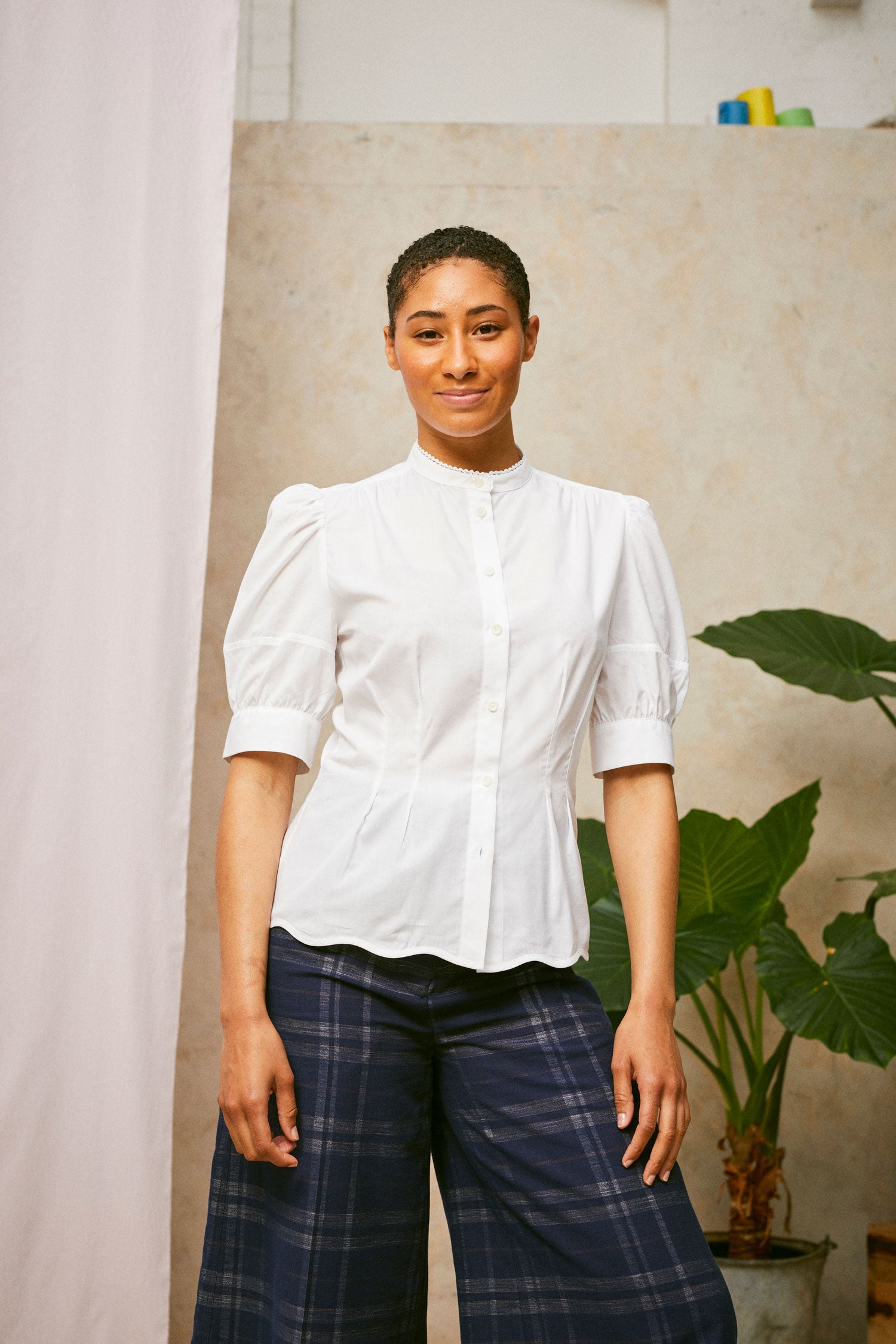Model stands smiling, wearing Saywood's Joni puff sleeve blouse in white with scalloped hem, worn with navy check Amelia wide leg trousers. A plant and drop of pink fabric can be seen in the background.