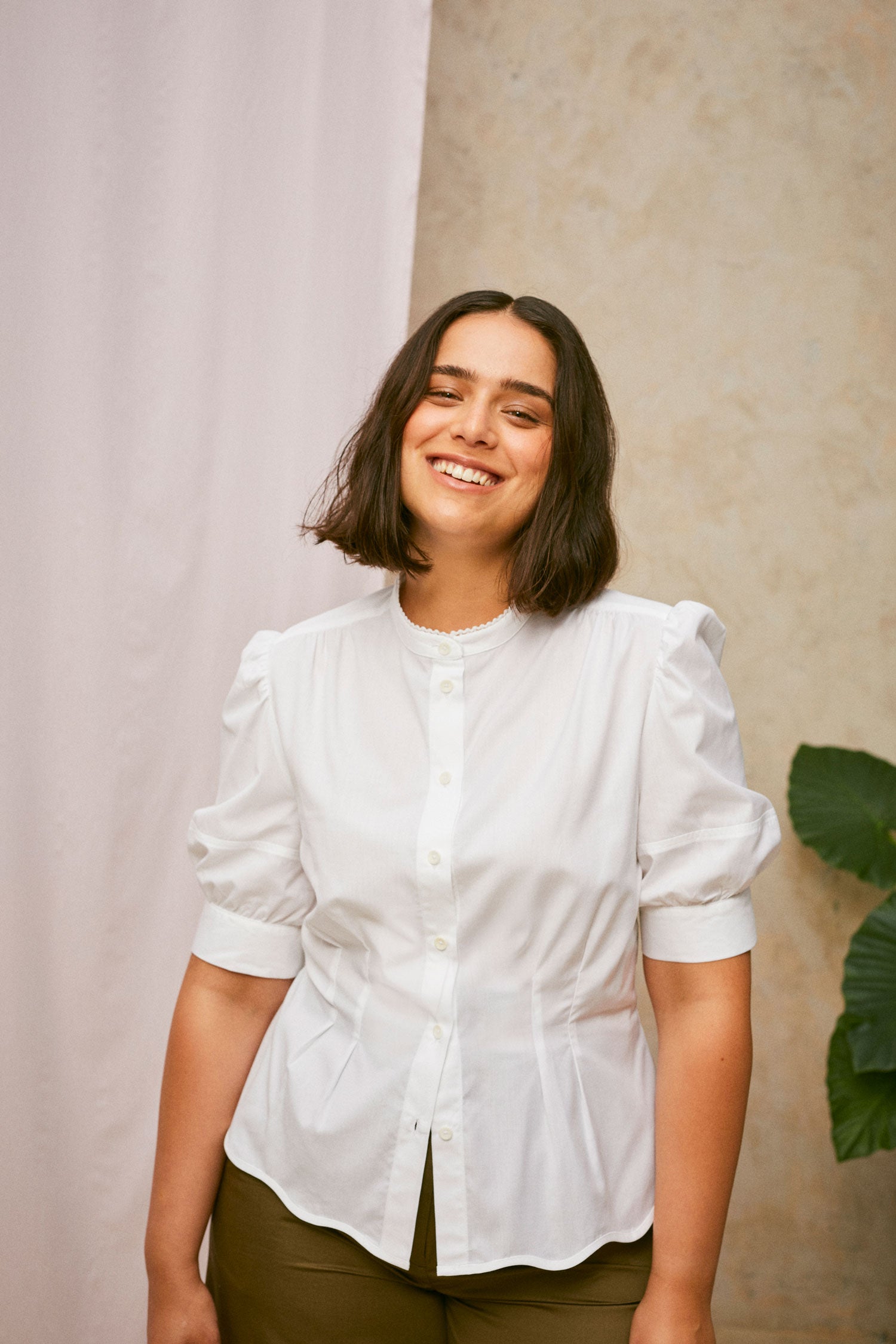 Model stands smiling, wearing Saywood's Joni puff sleeve blouse in white with scalloped hem, worn with khaki Amelia wide leg trousers. A plant and drop of pink fabric can be seen in the background.