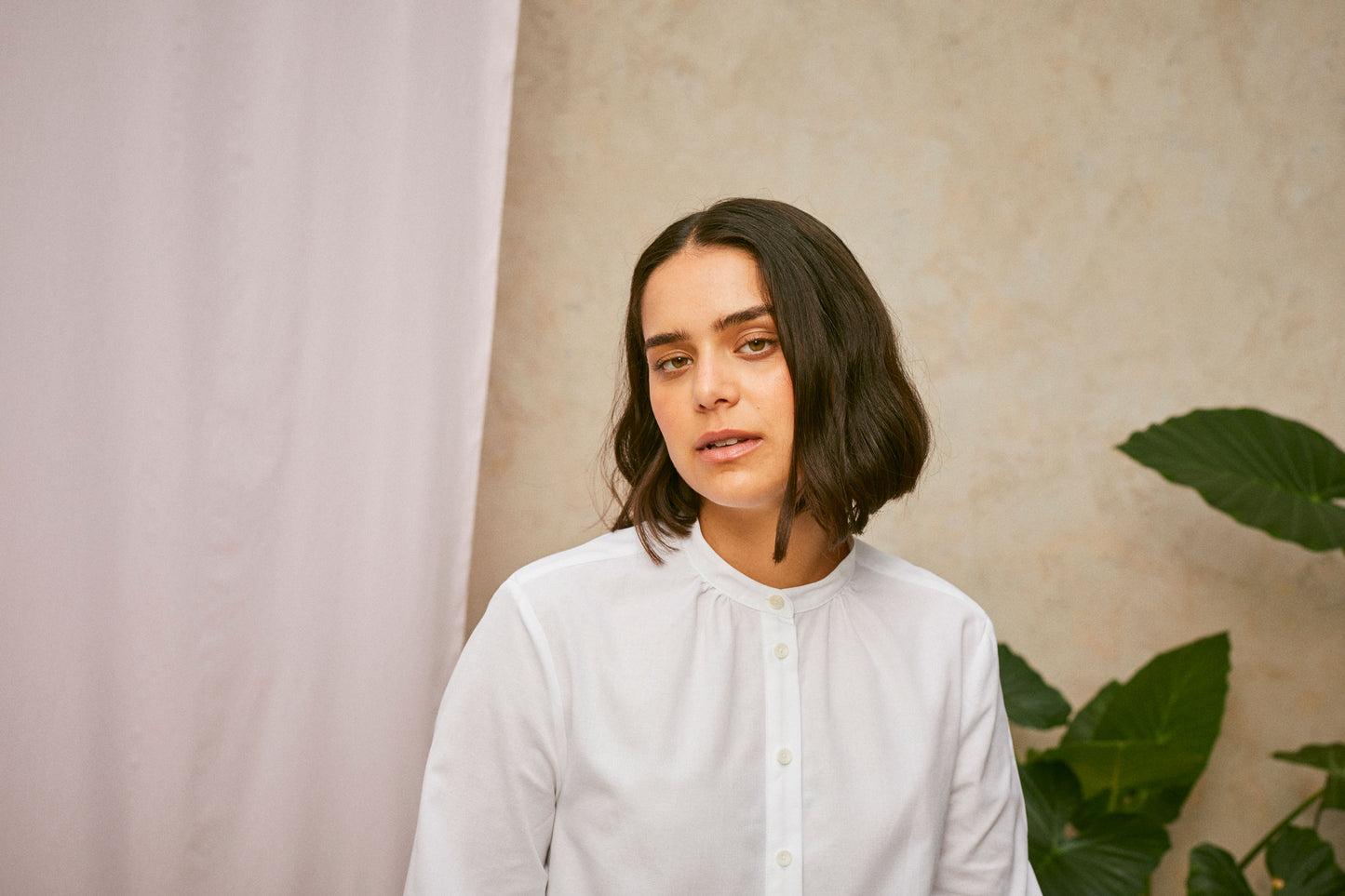 Close up of model wearing Saywood's white shirt, the Marie A-Line Blouse. The soft gathers at the neck can be seen. A plant and drop of pink fabric can be seen in the background.