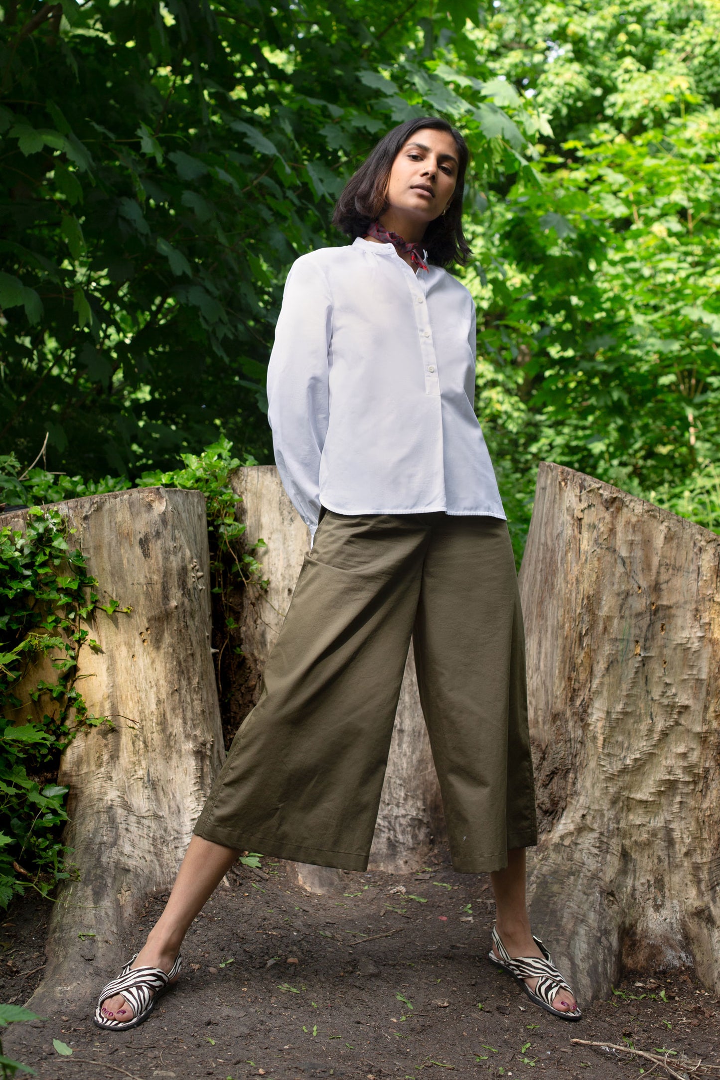 Model stands, legs in wide stance, in front of tree stumps and greenery. She wears Saywood's white Marie Aline Blouse with a red check neckerchief tied round her neck. Worn with Saywood's Amelia wide leg trouser in khaki and black and white zebra print sandals.