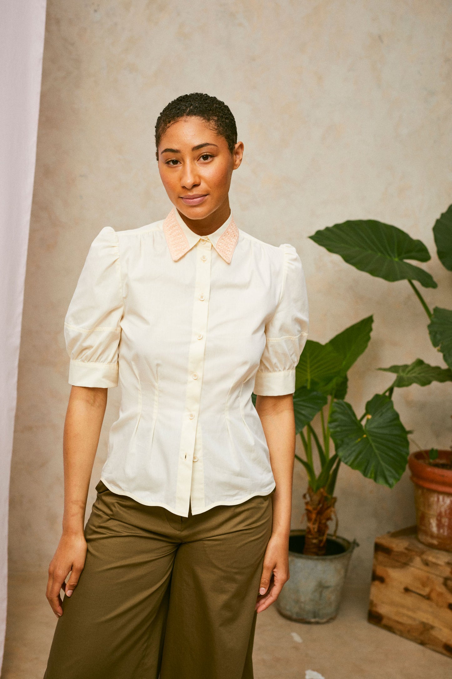 Model stands, wearing Saywood's pastel yellow Joni puff sleeve blouse with orange lace collar. Worn with khaki Amelia wide leg trousers. A plant and drop of pink fabric can be seen in the background. 