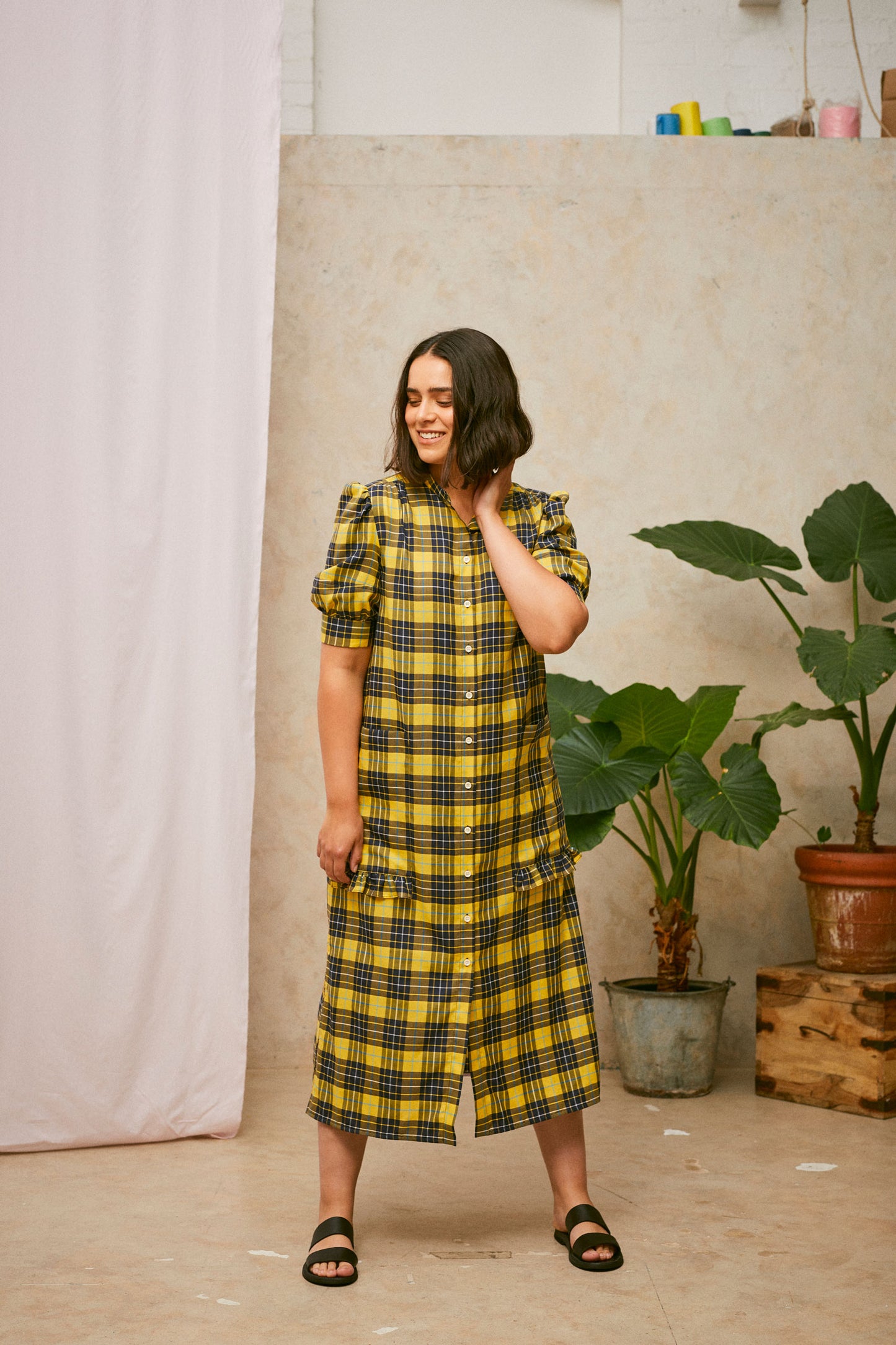 Full length shot of model wearing Saywood's Rosa yellow check shirtdress, loosely worn without a belt. Puff sleeves and patch pockets with ruffles are on the dress. Model has one hand to her neck and the dress is worn with black sandals. A plant and drop of pink fabric can be seen in the background.
